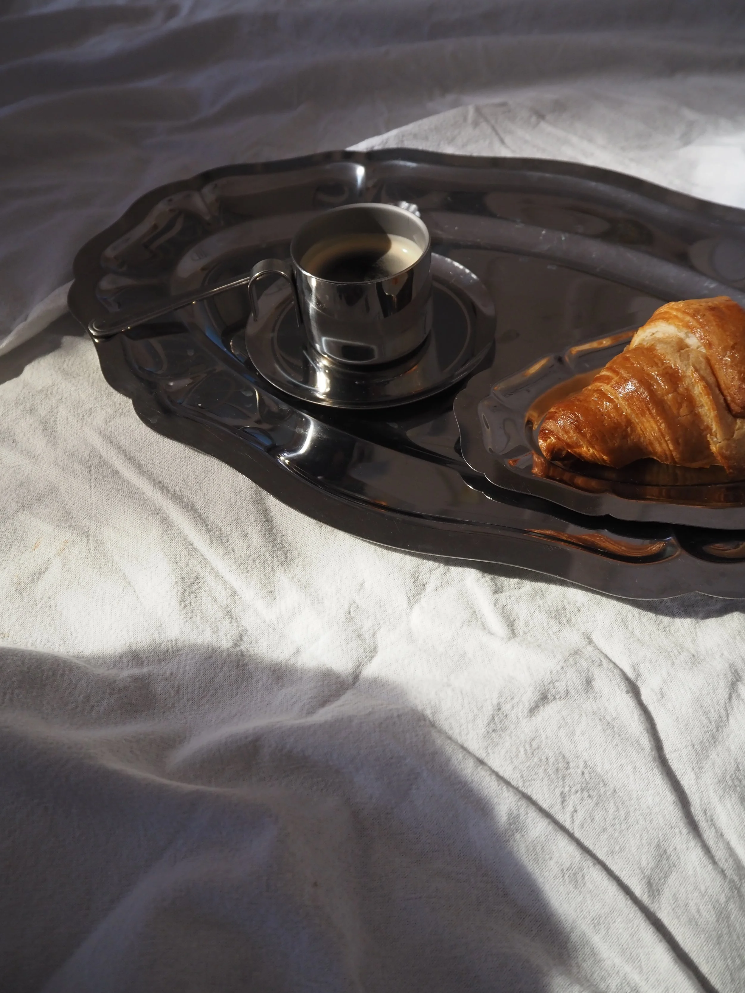 A silver tray holding a small cup of espresso coffee and a croissant on a white cloth surface.