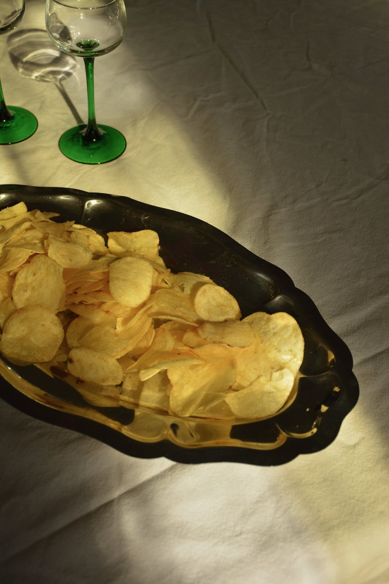 A black and gold dish filled with potato chips on a white tablecloth, with two green and clear wine glasses in the background.