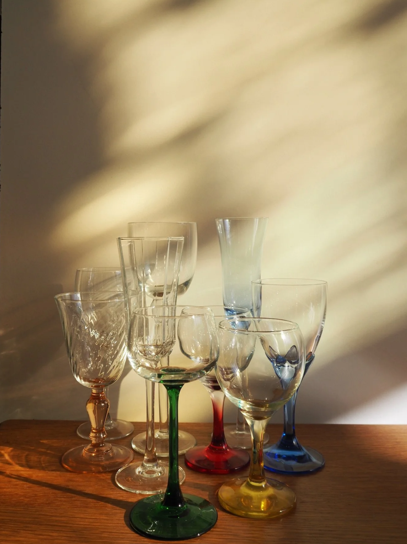A collection of nine colorful glassware glasses on a wooden surface, with a soft shadow cast on a beige wall behind them.