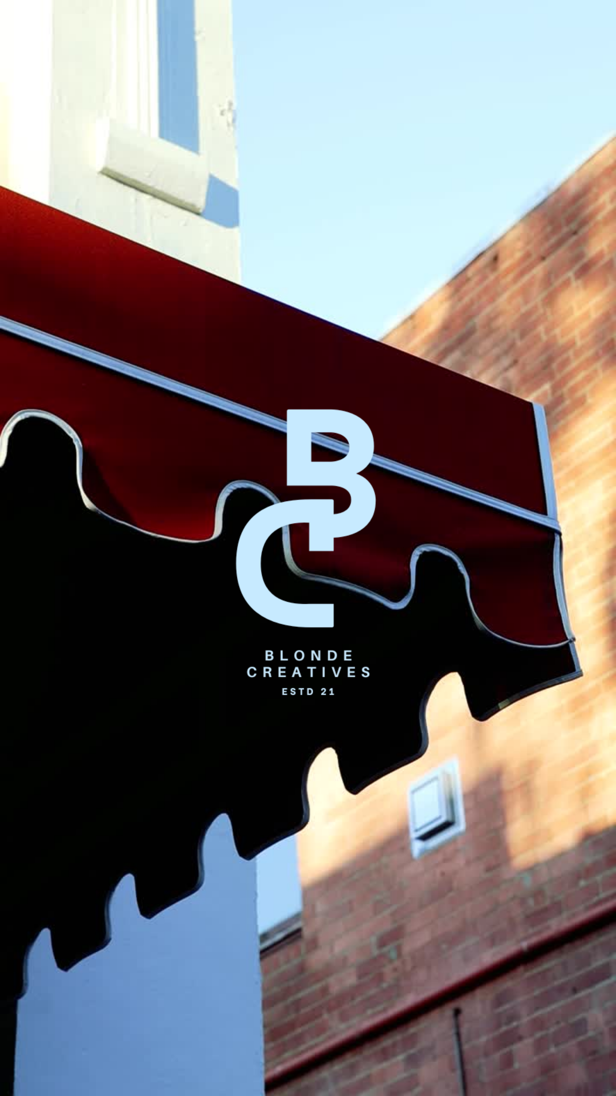 Blonde Creatives BC monogram logo displayed on a red and black storefront awning against a brick building exterior.