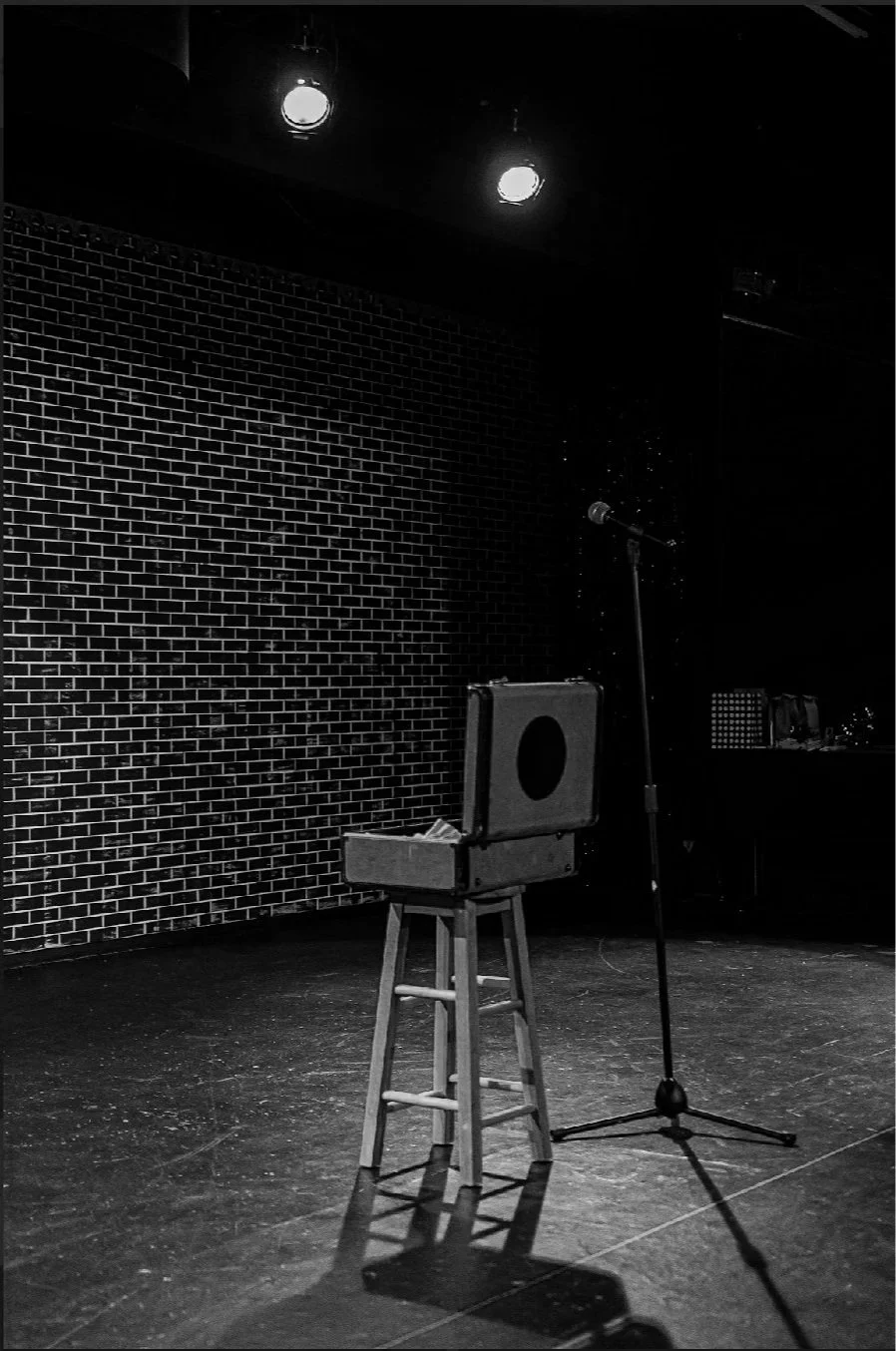 Empty comedy stage with a stool, an open box, and a microphone stand in front of a brick wall, under stage lights.
