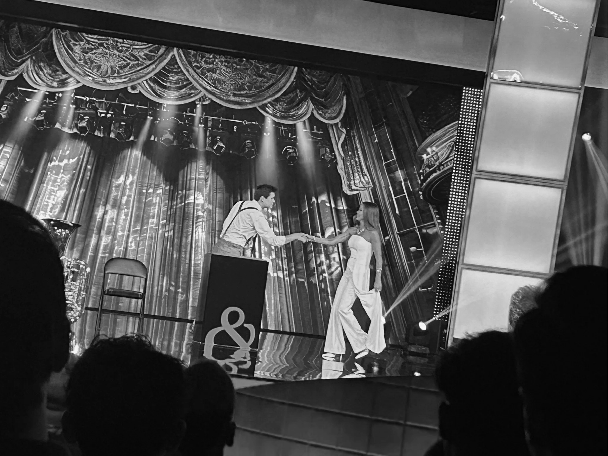 A black and white photo of a stage with two performers. One man is handing a flower to a woman. The stage has curtains and lighting, with an audience in the foreground.