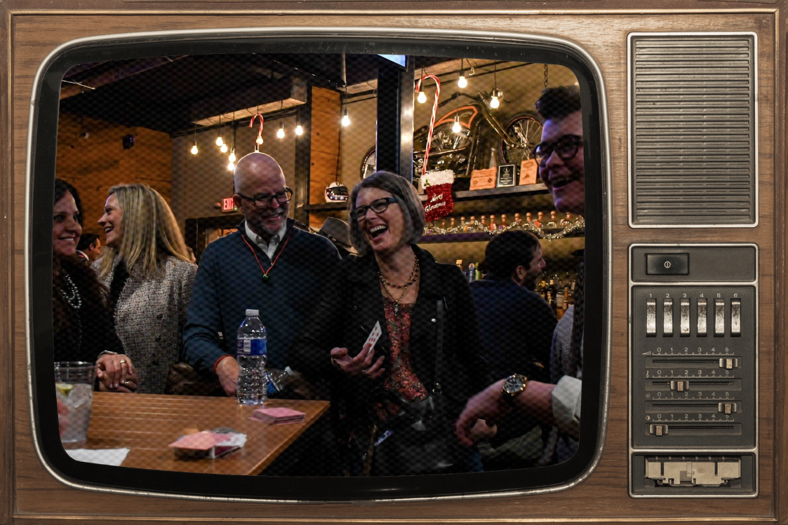 People at a bar or party viewed through a vintage television screen, laughing and talking, with Christmas decorations and a motorcycle in the background.