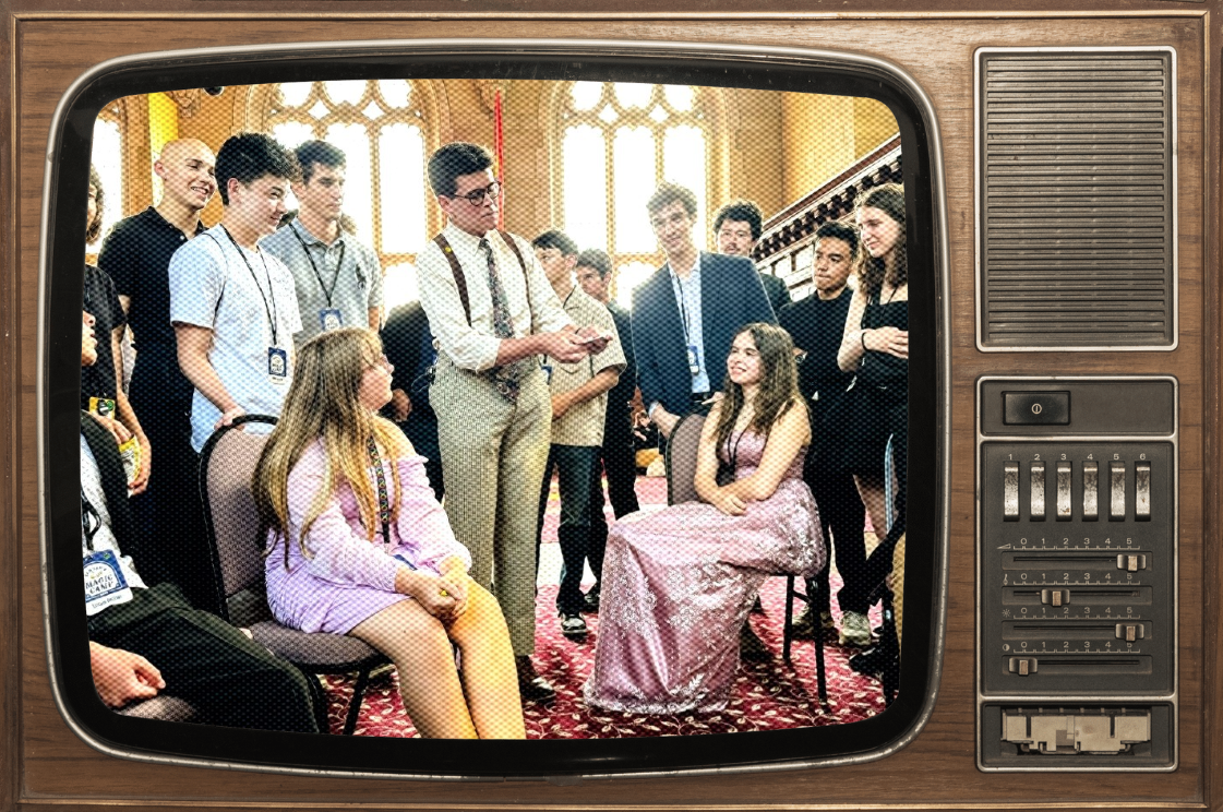 A vintage television screen displaying a scene with a group of people, including two young girls in dresses, gathered in an ornate room with large stained glass windows. Some people are sitting, some standing, and one man is holding a phone, appearing to be in a discussion or interview.