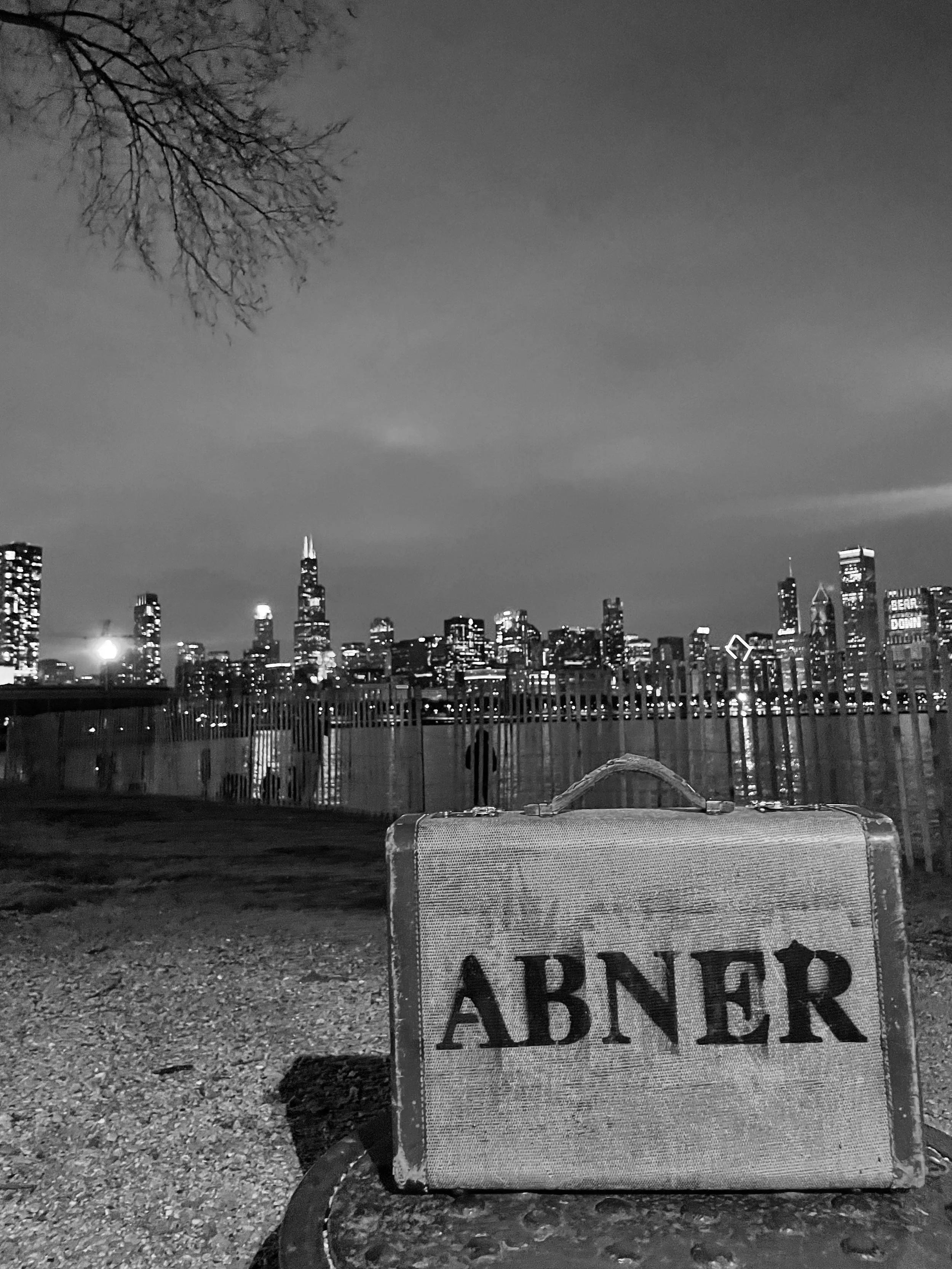 A vintage suitcase with the word 'ABNER' written on it, placed on the ground in front of a waterfront with Chicago's skyline in the background, at night.