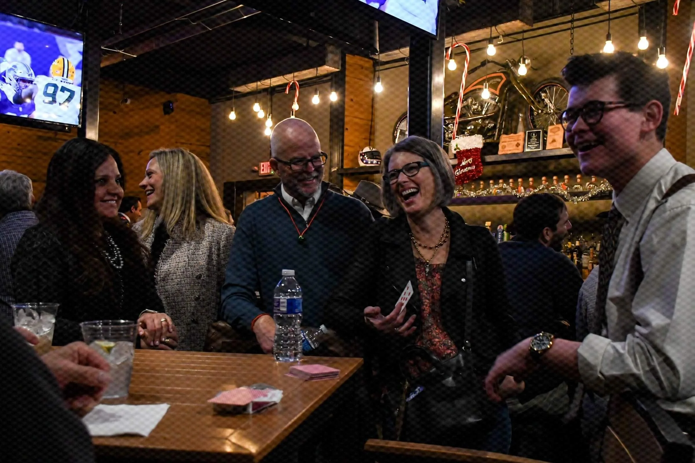 People laughing and talking at a social gathering in a warmly lit restaurant or bar with a large motorcycle decoration and Christmas garland in the background.