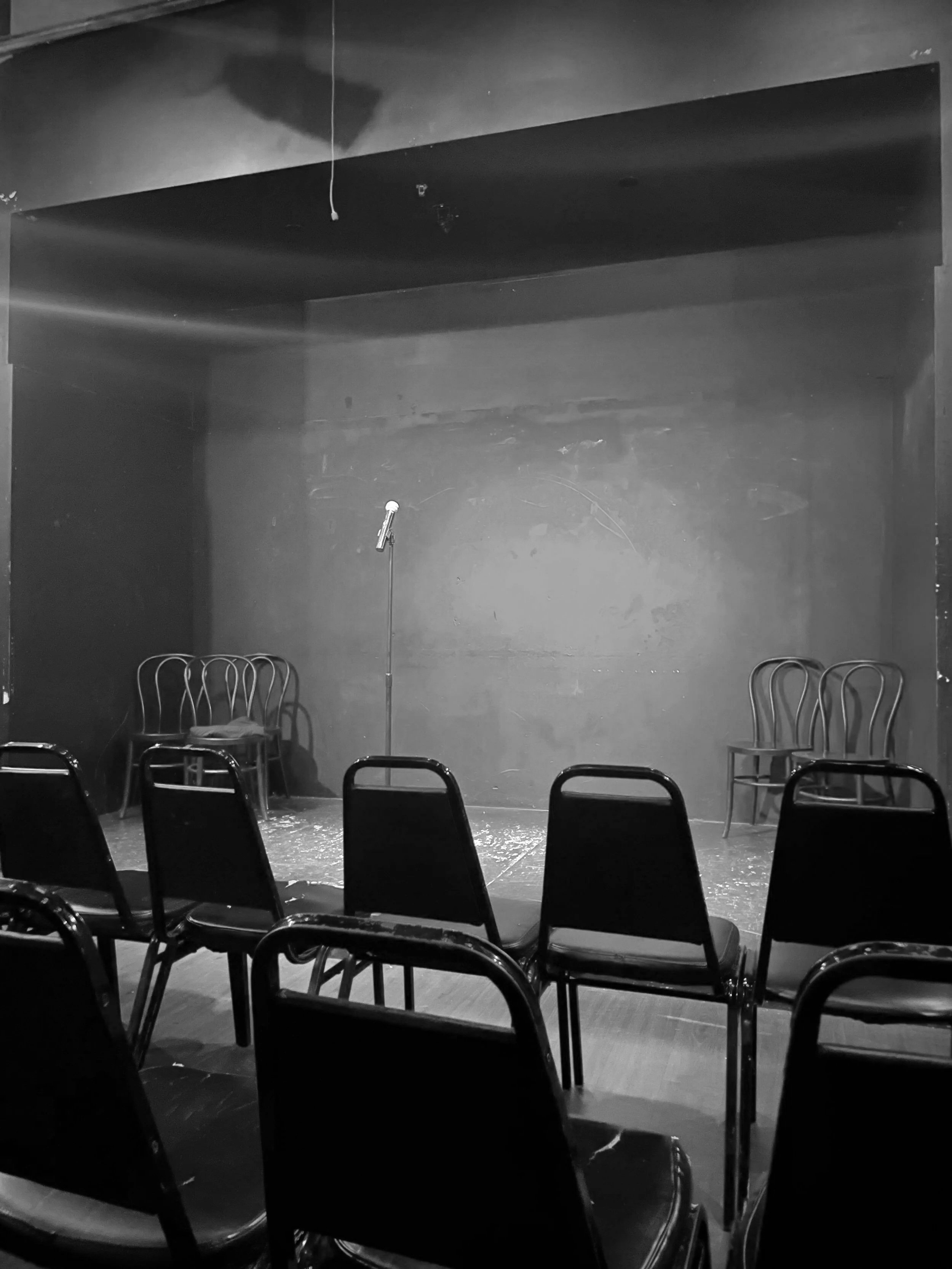 Empty stage with six chairs, a microphone on a stand, and a dark wall backdrop in a black-and-white photo.