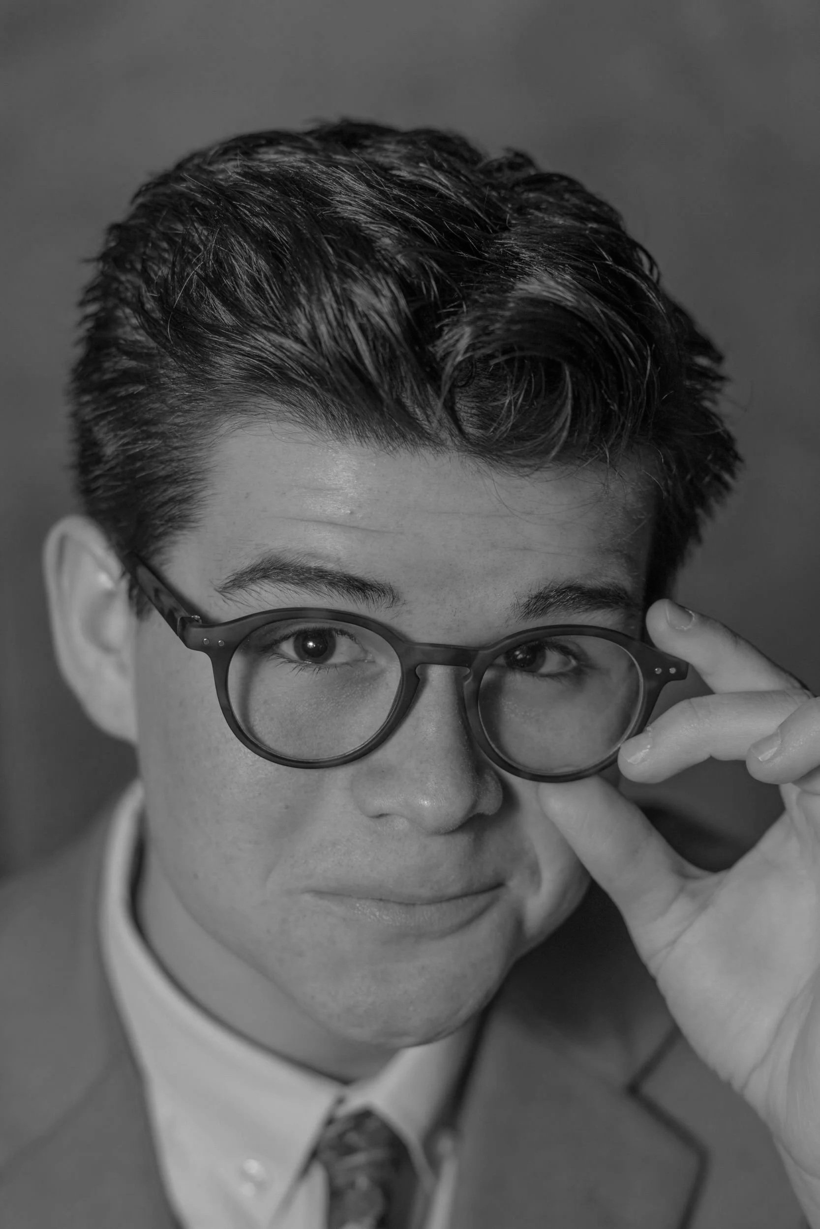 Black and white portrait of a young man with neatly styled hair, wearing glasses, and adjusting his glasses with one hand, smiling subtly, dressed in a suit and tie.