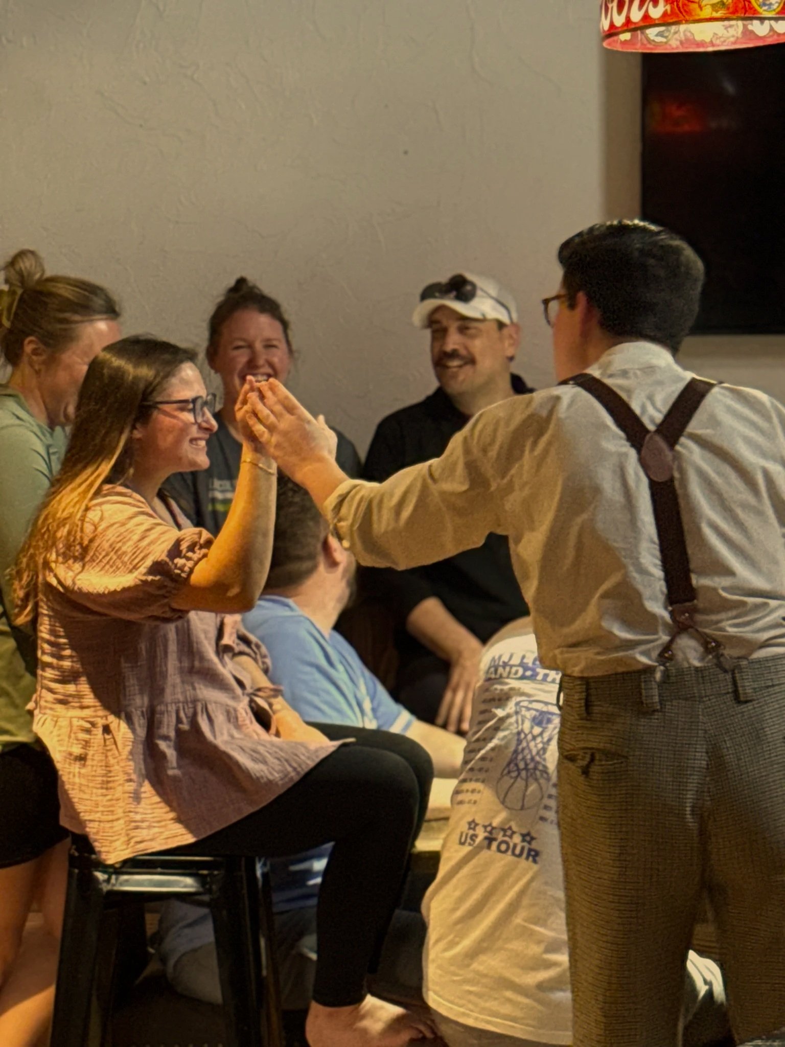 A group of people gathered indoors, laughing and engaging in conversation, with one person sitting and others standing or leaning nearby.
