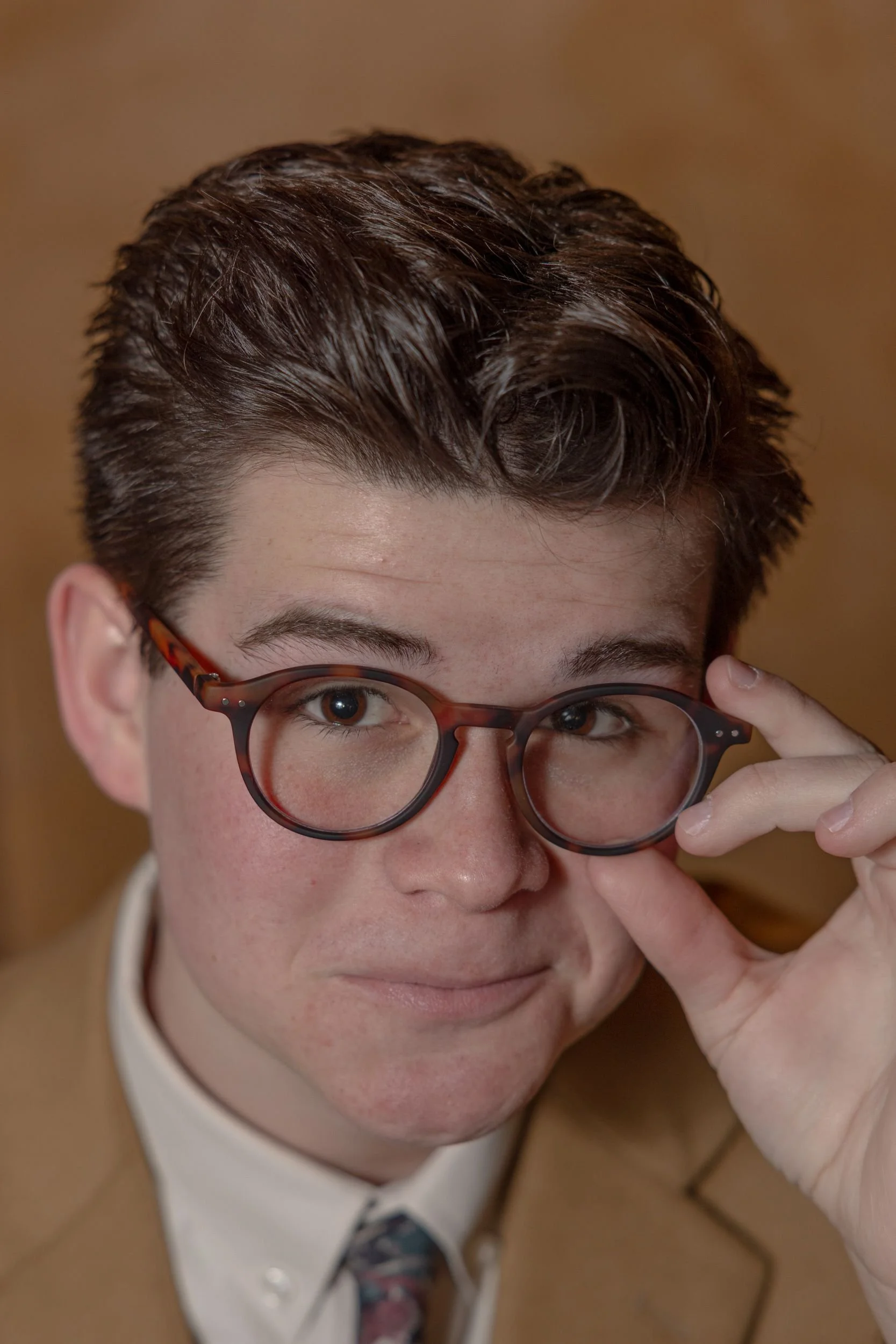 A young man with dark, wavy hair wearing tortoiseshell glasses and a beige blazer, adjusting his glasses with his hand, looking into the camera.