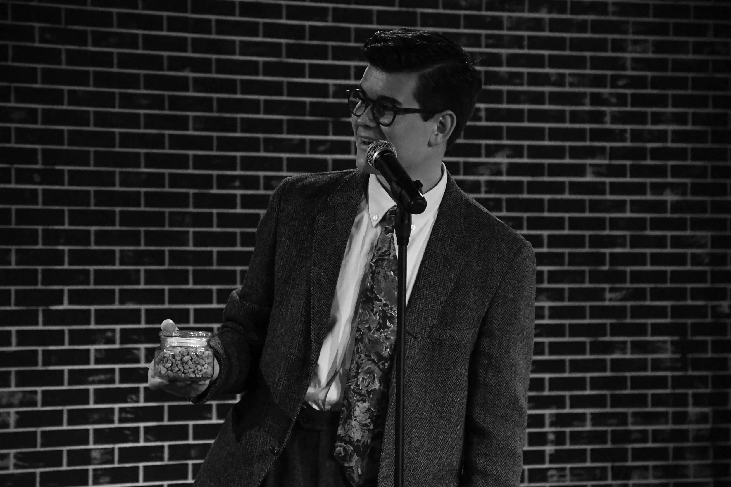 A young man with glasses, wearing a suit jacket and a patterned shirt, stands in front of a brick wall while speaking into a microphone. He holds a jar filled with candies in his left hand.