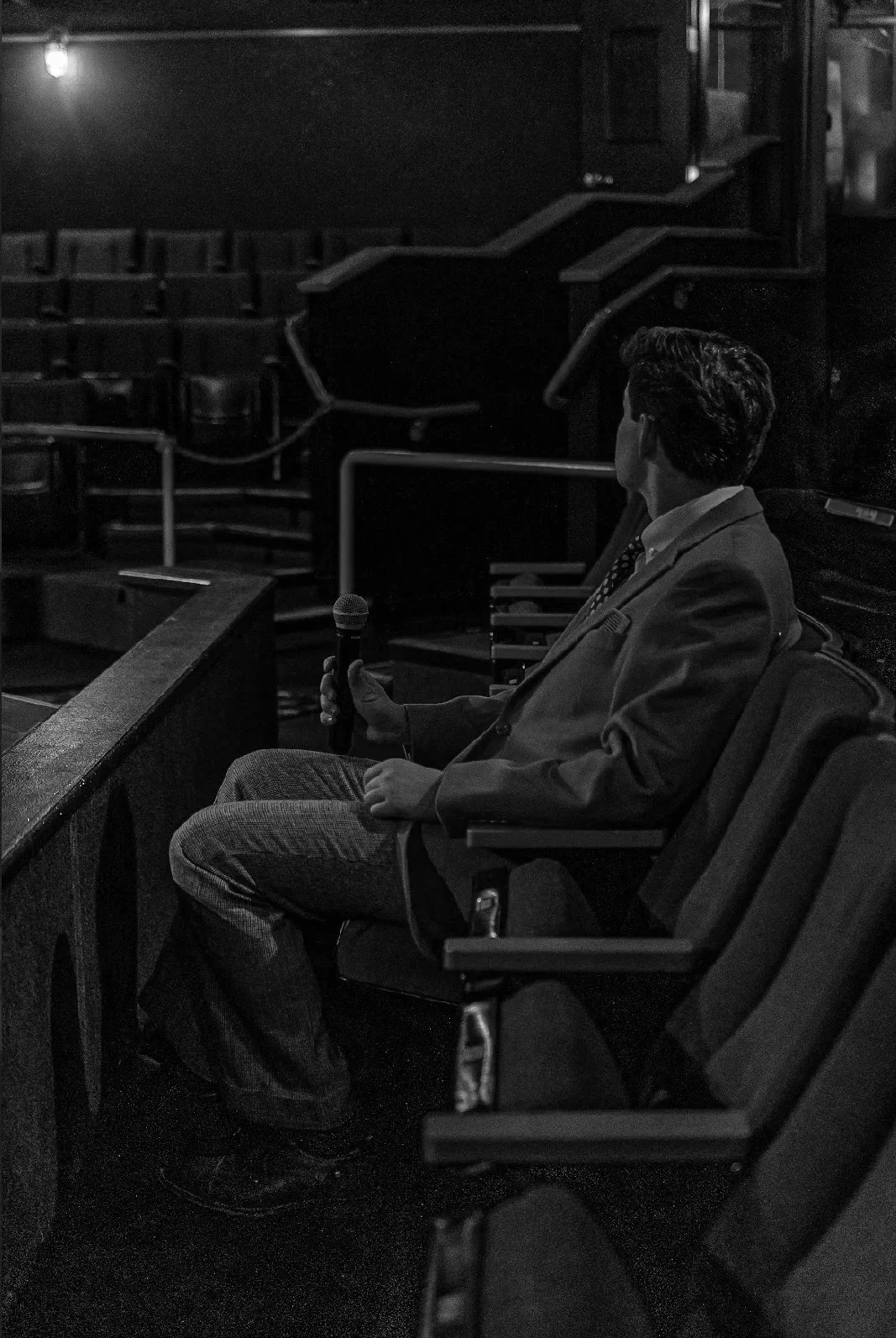 A man in a suit sitting alone in an auditorium, holding a microphone in his right hand, facing away from the camera.