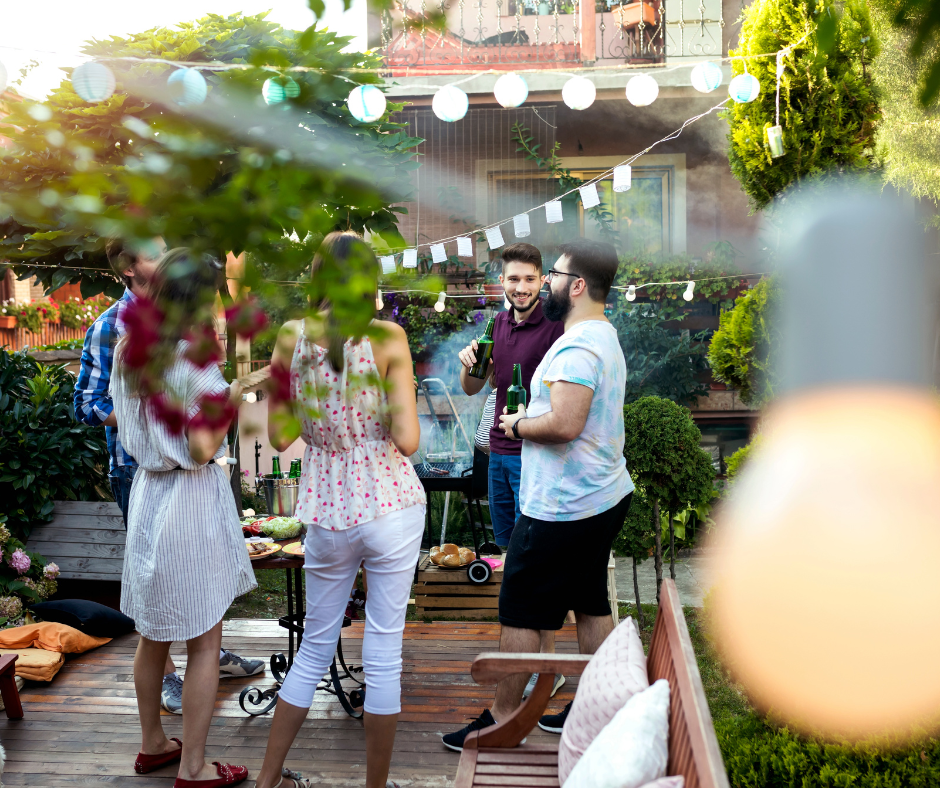 Group of friends chatting and drinking beers on a backyard patio decorated with string lights and greenery.