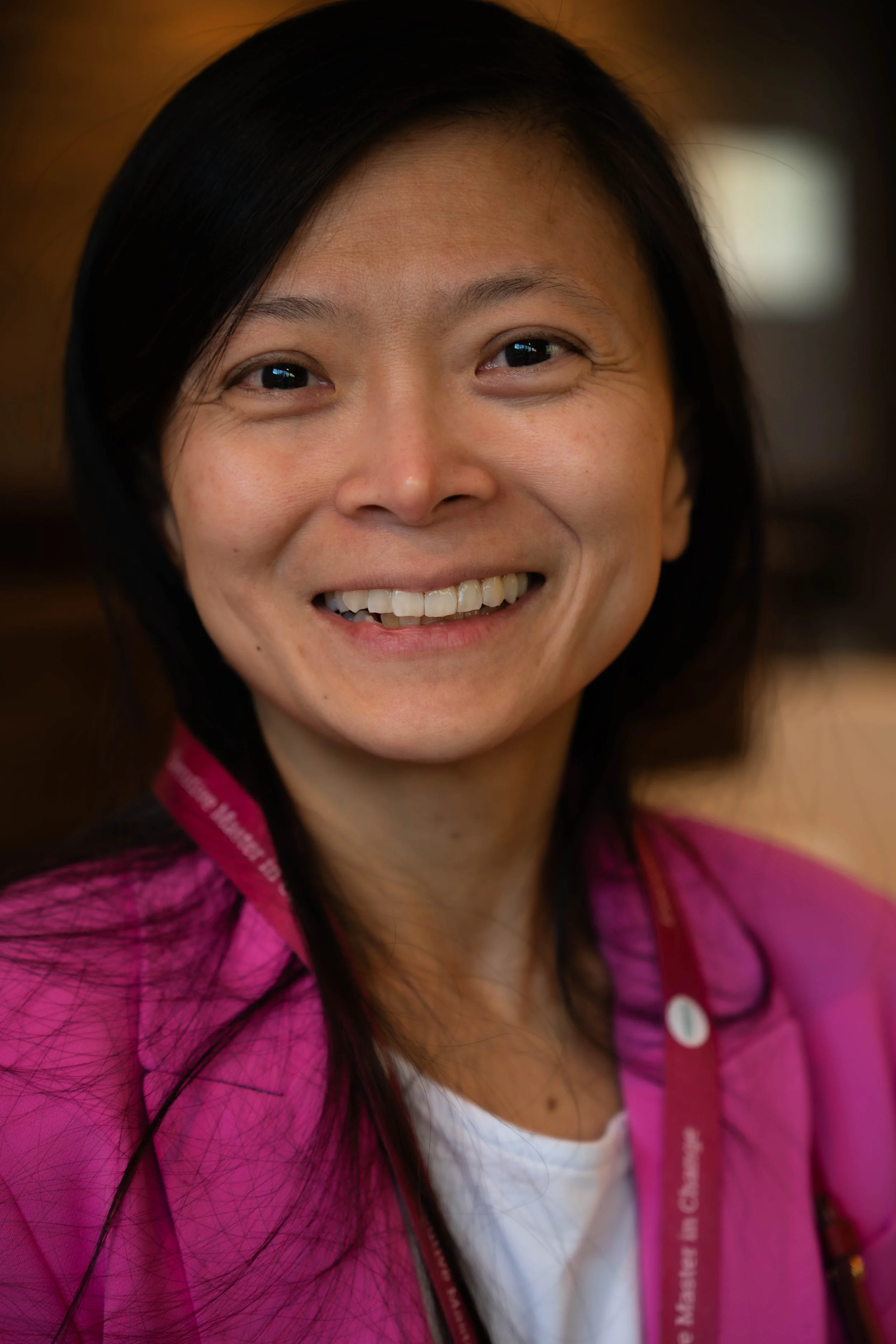 Close-up of a smiling woman with black hair, wearing a pink jacket and a white shirt.