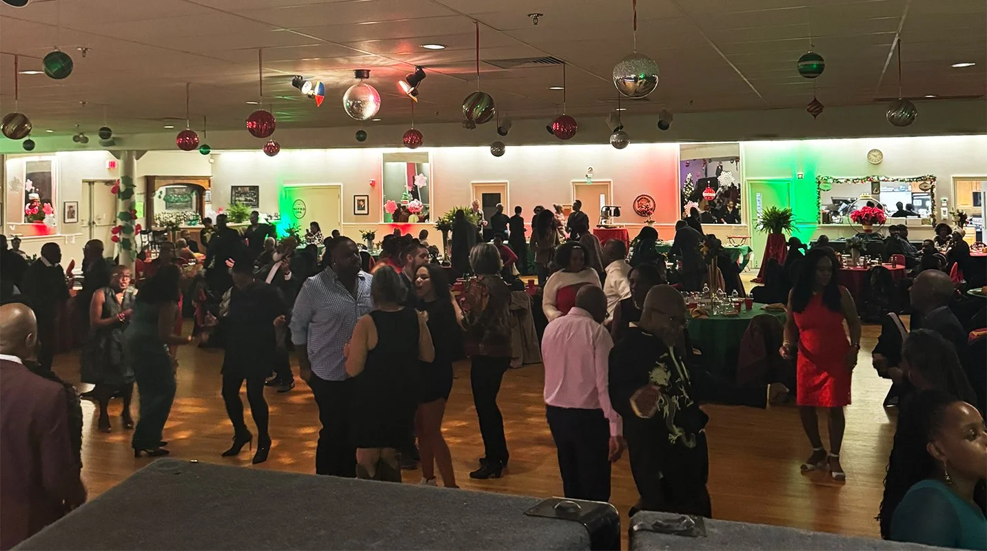 People dancing and socializing at a festive celebration in a decorated hall with Christmas ornaments and colorful lighting.