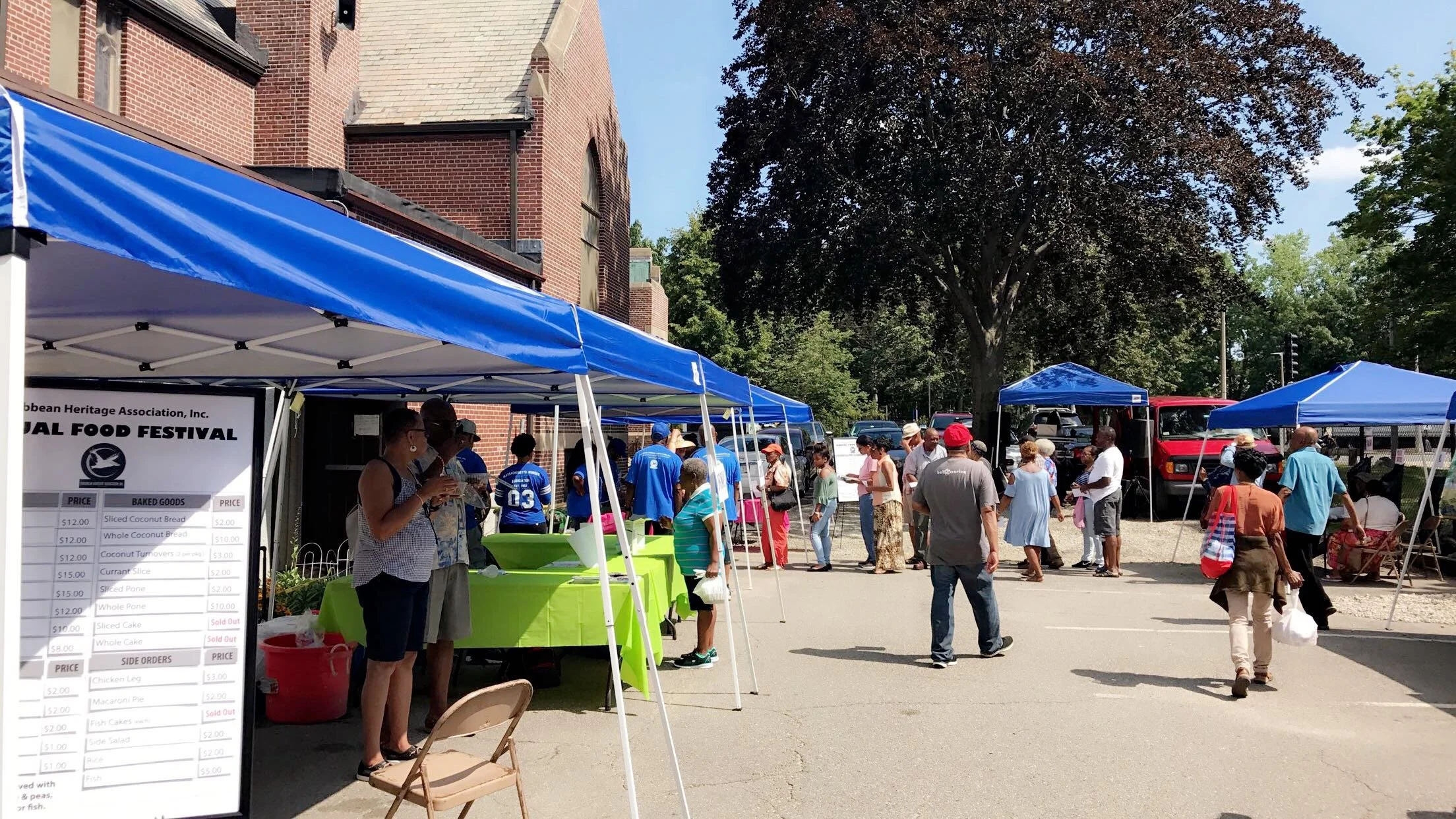 People browsing and purchasing at an outdoor food festival with blue tents, a large tree, and a red brick building in the background.