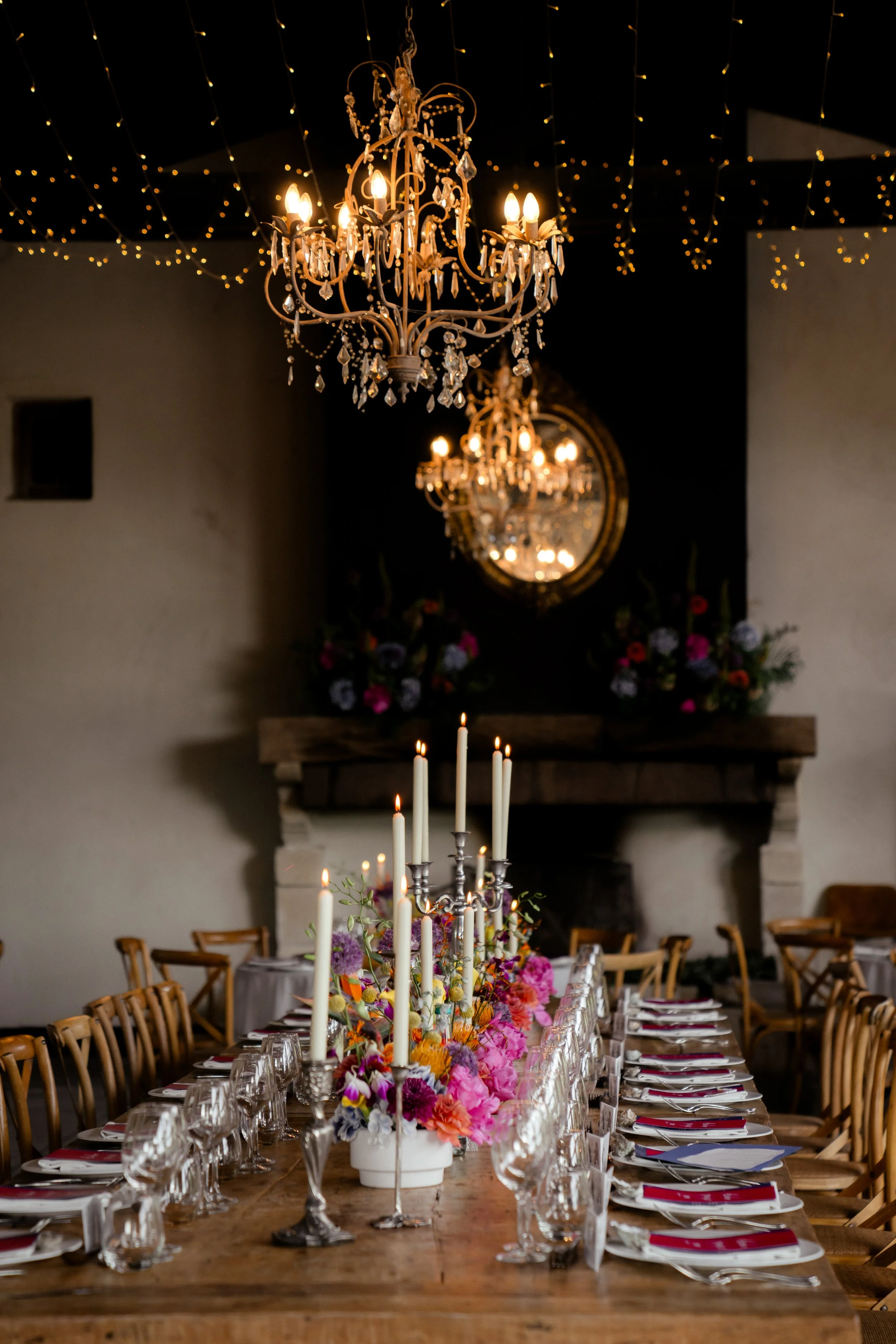 Ein festlich gedeckter Tisch mit Kerzen, Blumen und Kristallgläsern in einem eleganten Raum mit Kronleuchtern und Dekor wie Lichterketten.