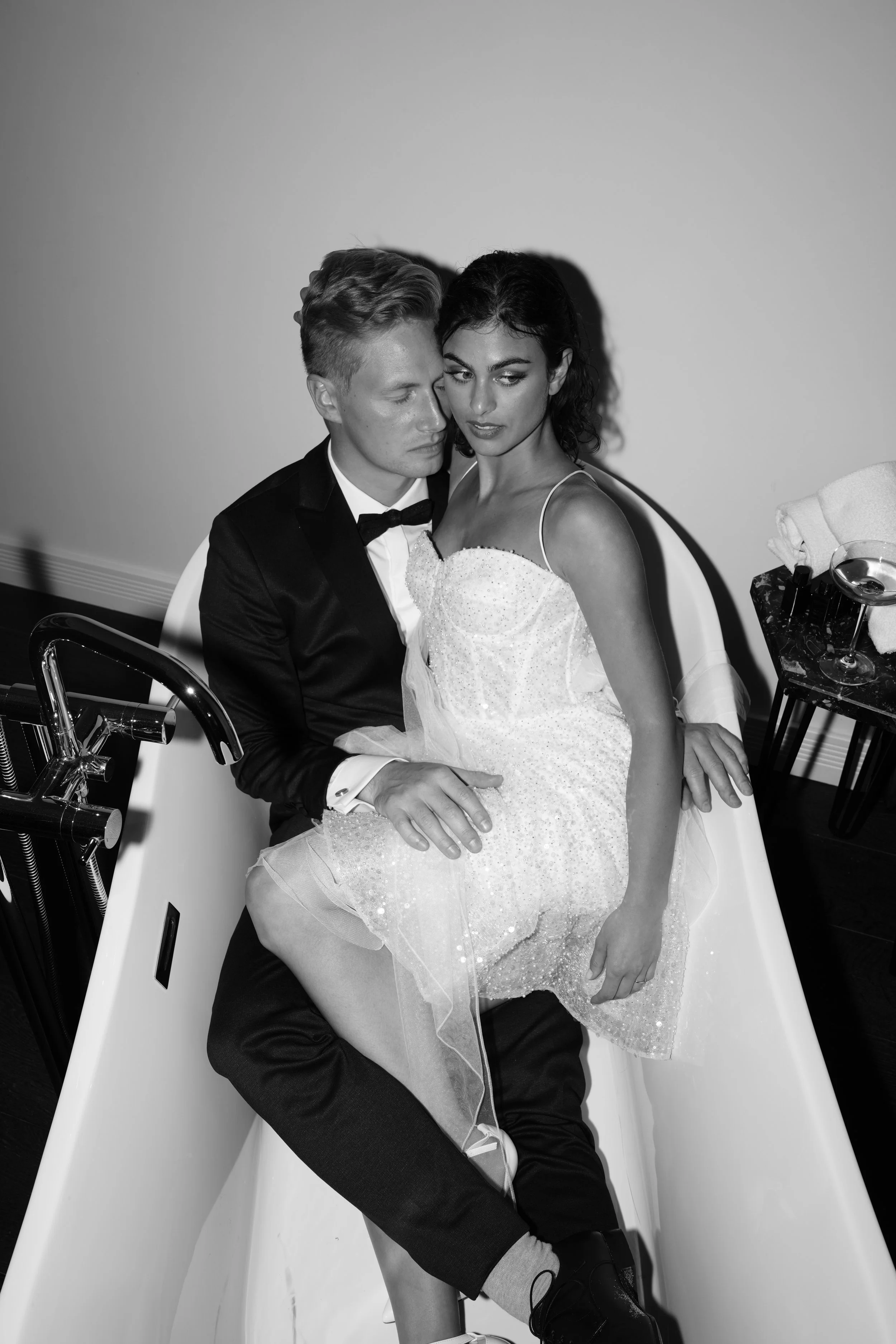 Ein Mann und eine Frau sitzen in einer Badewanne, die mit Wasser gefüllt ist. Der Mann trägt einen schwarzen Anzug mit Fliege, die Frau trägt ein weißes Kleid. Es ist ein Schwarz-Weiß-Foto.