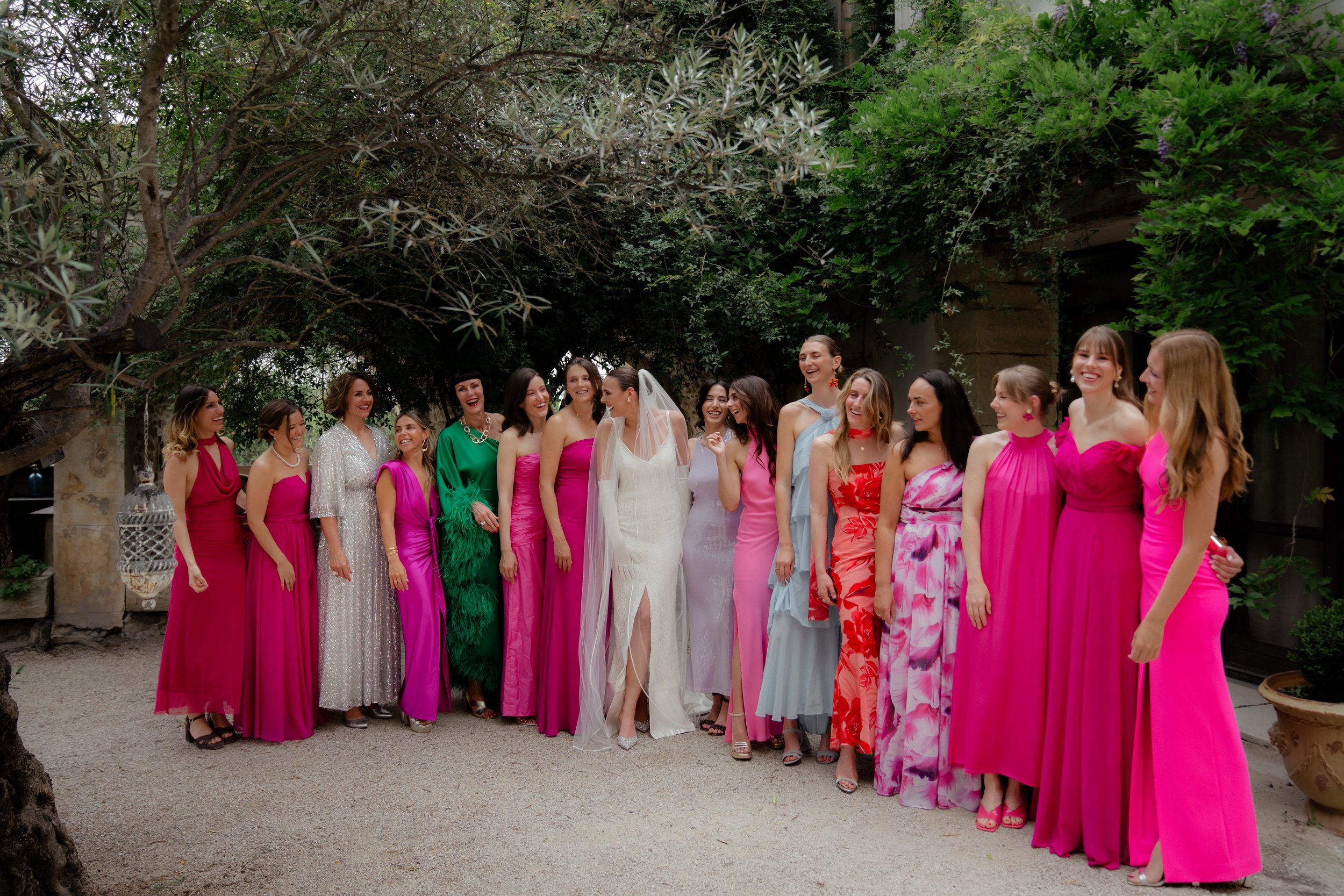 Gruppe von Frauen, darunter eine Braut in weißem Kleid mit Schleier, in einem Garten mit viel Grün, lachen und unterhalten sich, alle tragen bunte Kleider in Pink, Rot, Blau und Grün.