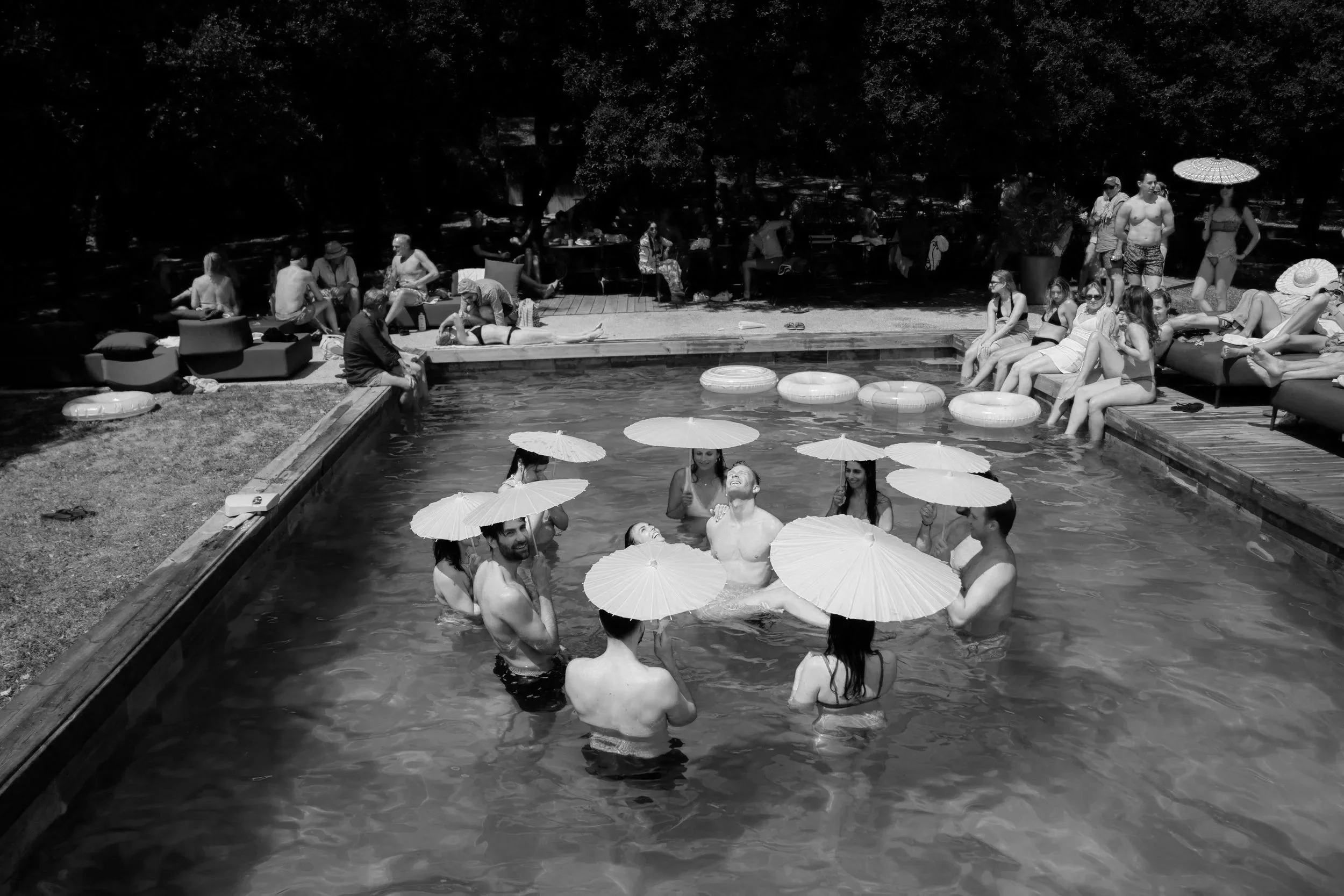 Menschen in einem Pool, die mit weißen Papier-Schirmen Sonnenbaden gestalten, während andere am Poolrand entspannen und im Schatten sitzen, umgeben von Wasserauflegern