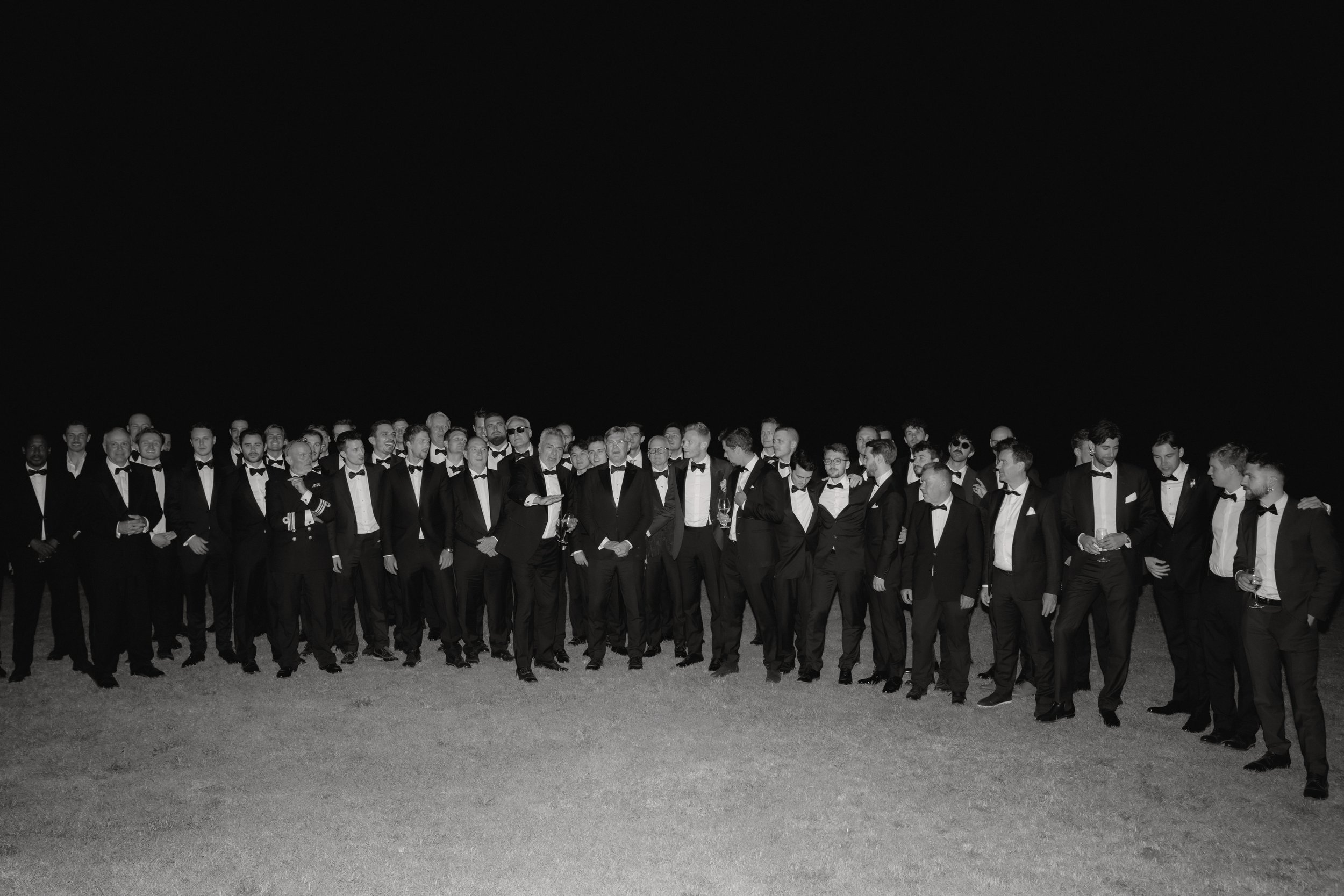 Gruppe von Männern in Smoking, die an einem Abend-Event teilnehmen, aufgenommen bei Nacht auf freiem Feld.
