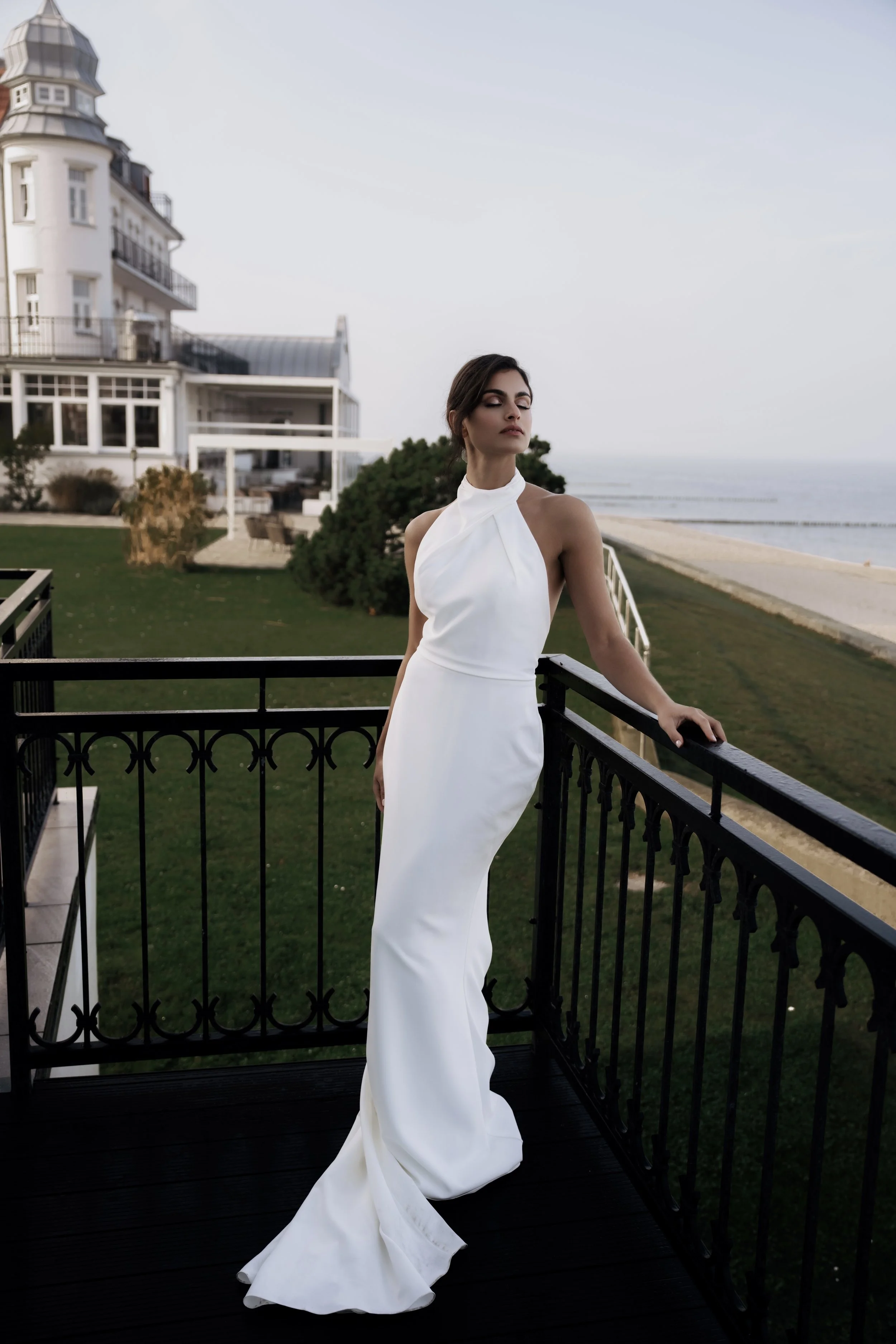 Frau in weißem Abendkleid steht auf einem Balkon mit Meerblick, vor einem großen weißen Haus.