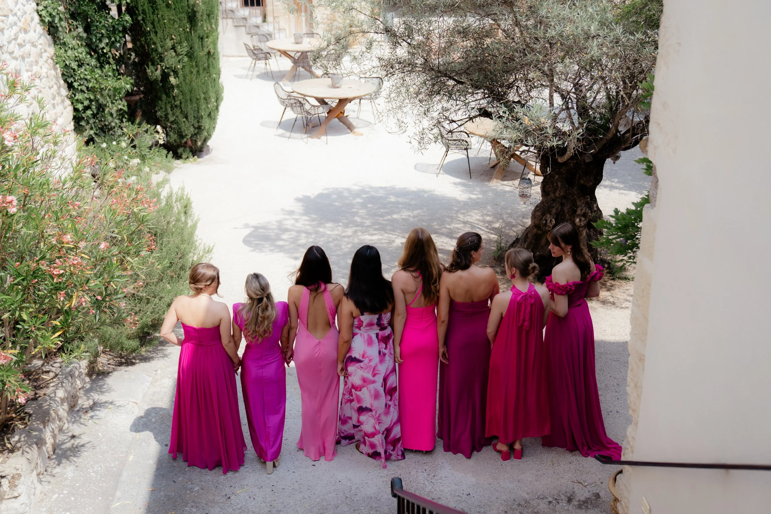 Eine Gruppe von Frauen in pinkfarbenen langen Kleidern steht in einer Reihe und hält sich an den Händen, im Freien vor einem Baum.