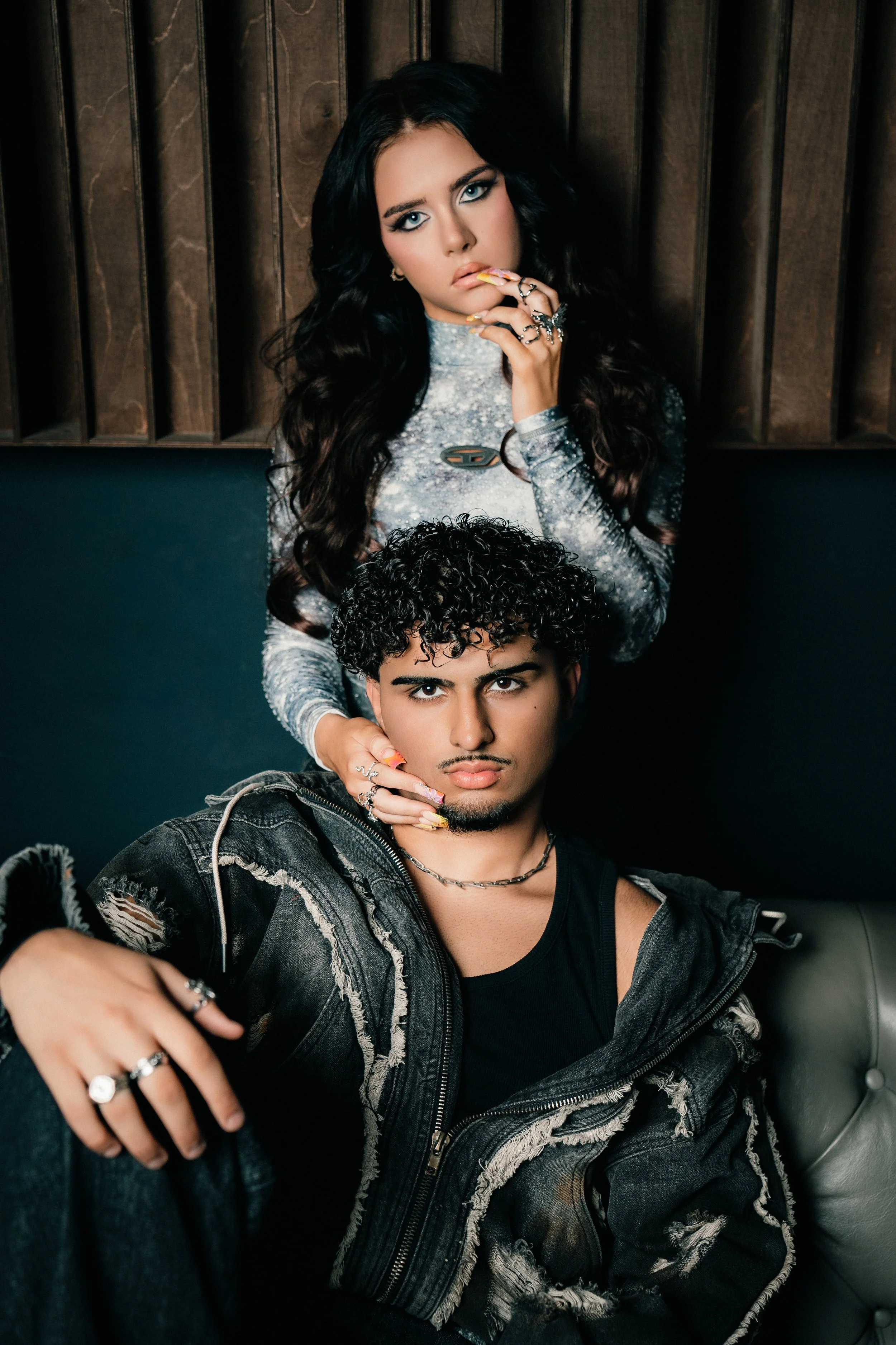 Ein Mann mit curly Haaren und schwarzen T-Shirt sitzt auf einem Sofa, während eine Frau mit langen dunklen Haaren und einem silbernen Oberteil hinter ihm steht, die ihn berührt.