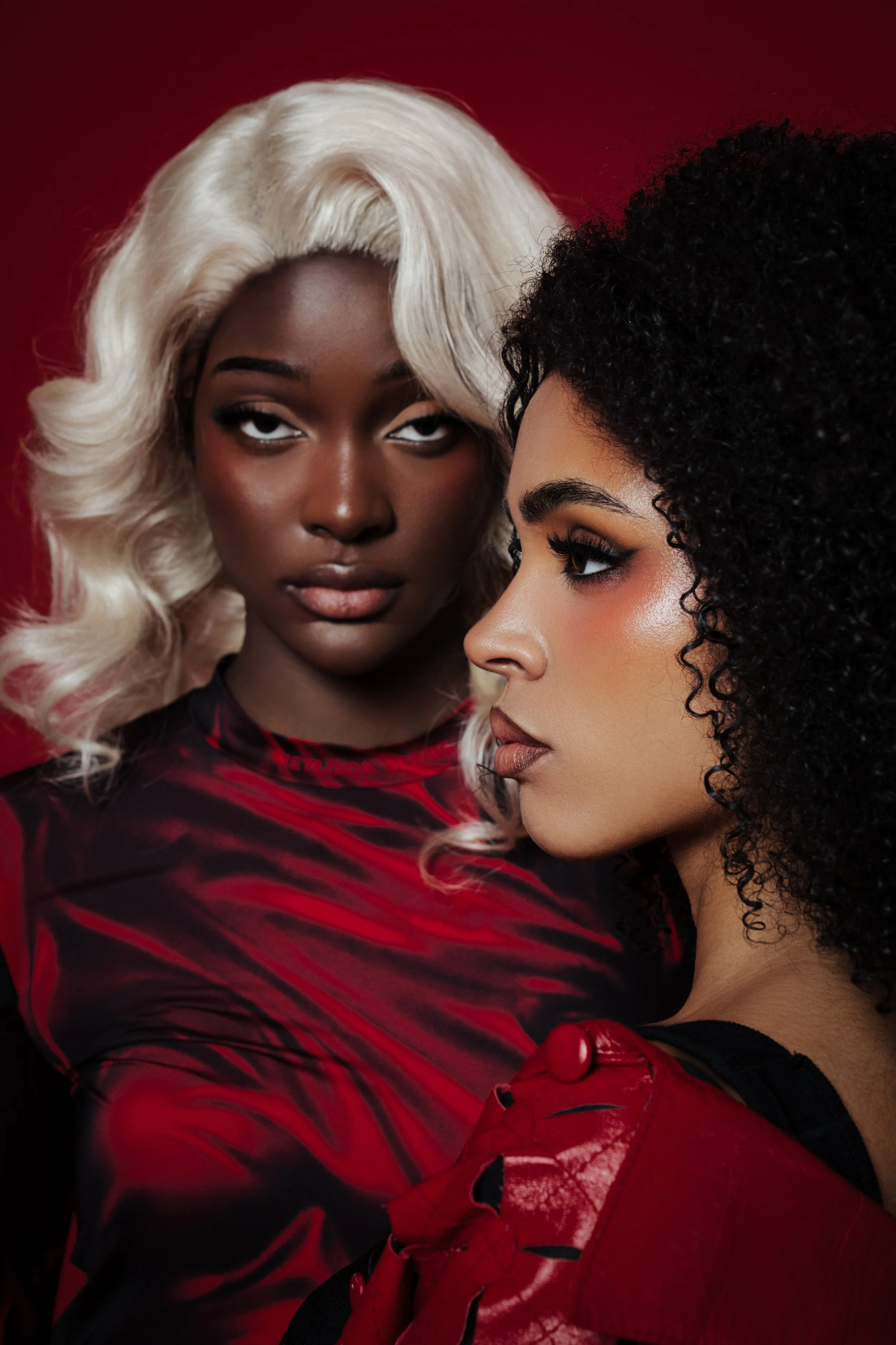 Zwei junge Frauen mit genauen Frisuren und Make-up vor rotem Hintergrund, eine mit platinblonden Locken und die andere mit schwarzen Locken, tragen schwarze und rote Kleidung.
