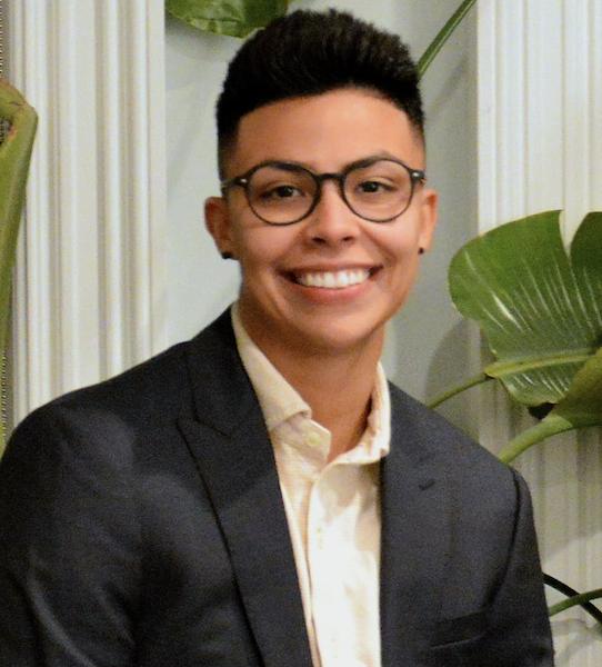 A smiling person with short dark hair, wearing glasses, a dark blazer, and a light-colored shirt, standing indoors near large green leafy plants.