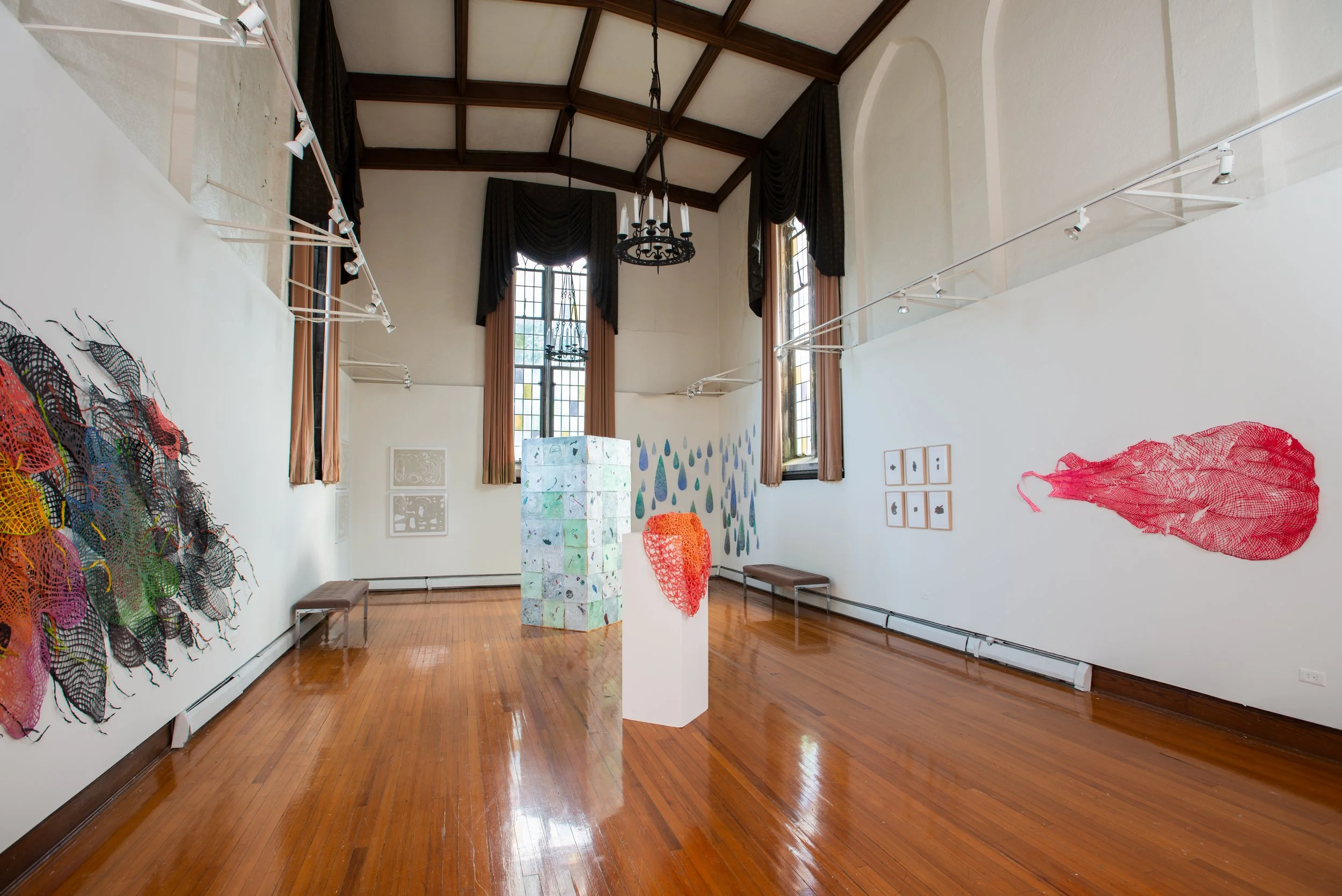 An art gallery with white walls, wooden floors, tall windows with brown curtains, and various colorful artworks on display including a large textured piece on the left, a cluster of white boxes with painted designs in the center, and conceptual art o