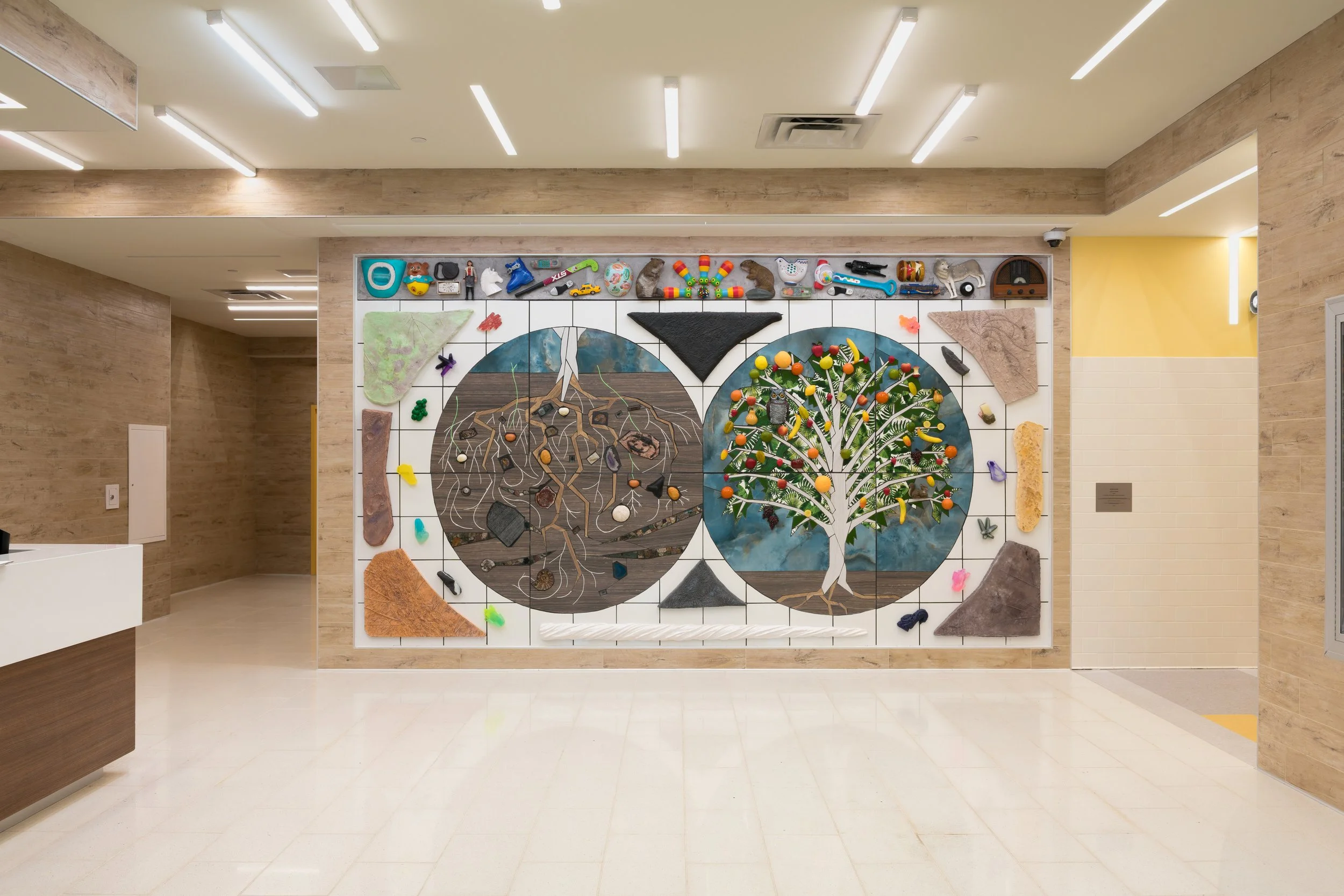 Colorful mosaic art installation on a wall in an indoor space depicting a tree and a connected underground root system, surrounded by various small objects and materials.