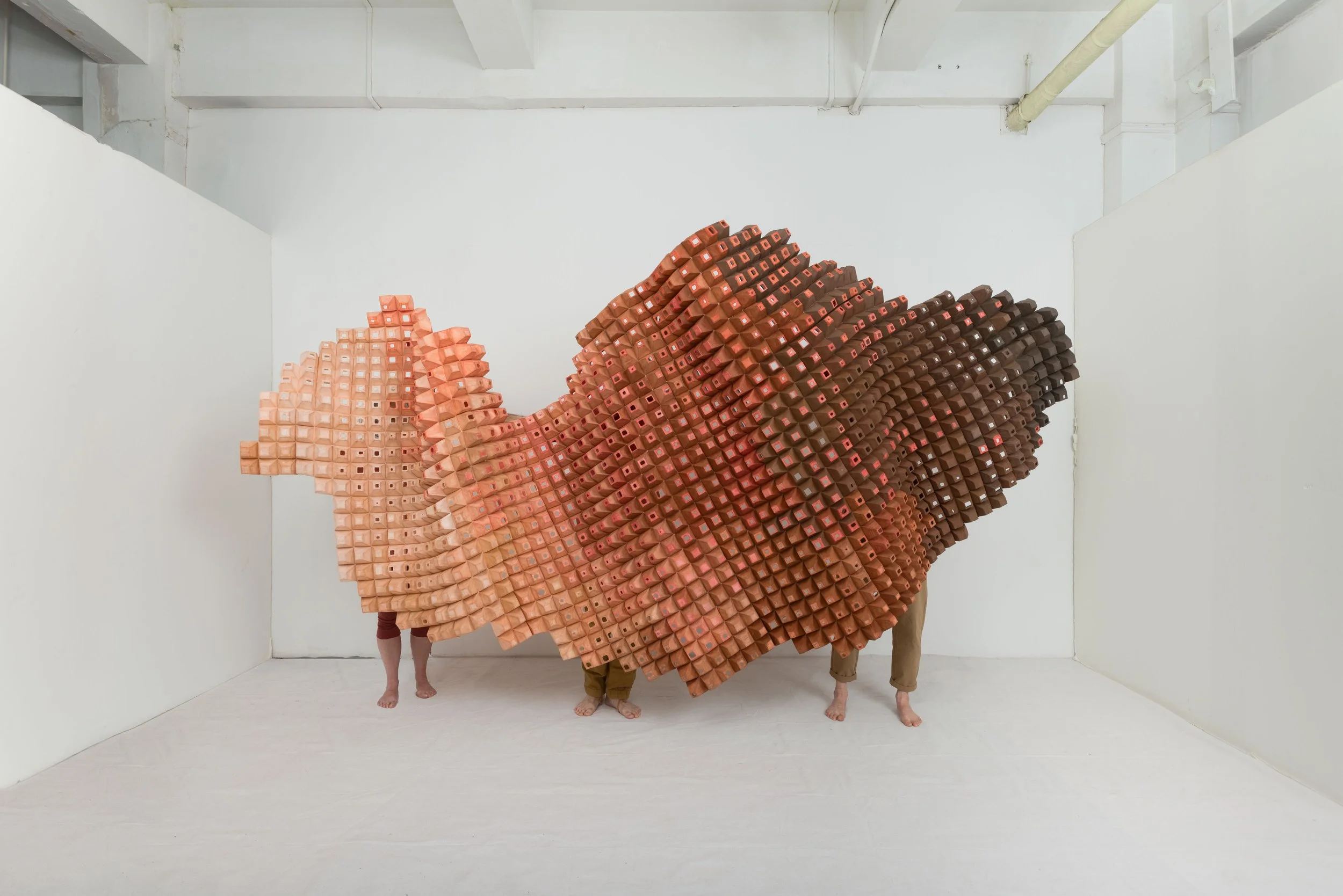 People partially obscured by a large sculpture resembling a fish, made of numerous small clay or ceramic blocks in shades of pink, orange, and brown, in a white-walled gallery space.
