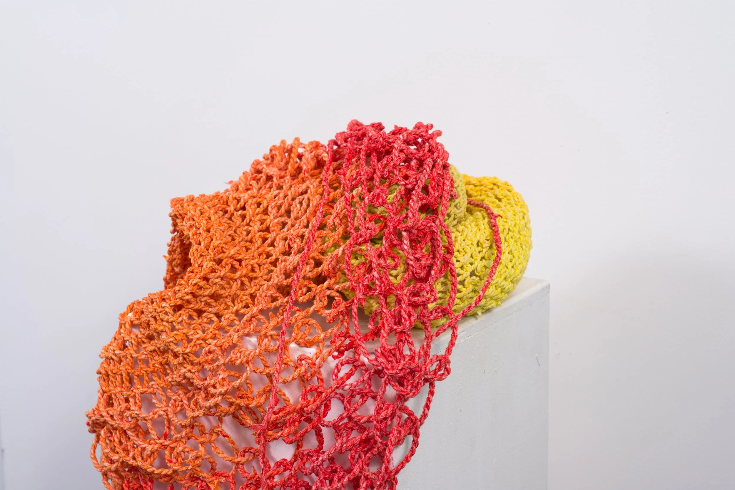 Colorful handmade crochet scarves in orange, pink, and yellow draped over a white pedestal against a plain white background.