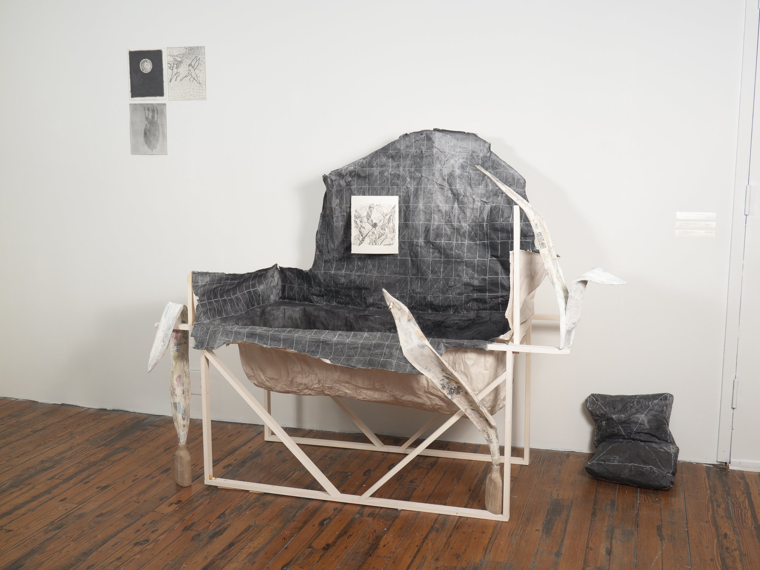 Mixed media art installation resembling a deconstructed sofa with black and gray fabric, paper, and wood, positioned on a wooden floor against a white wall, with small black and white images and sketches attached to the wall.