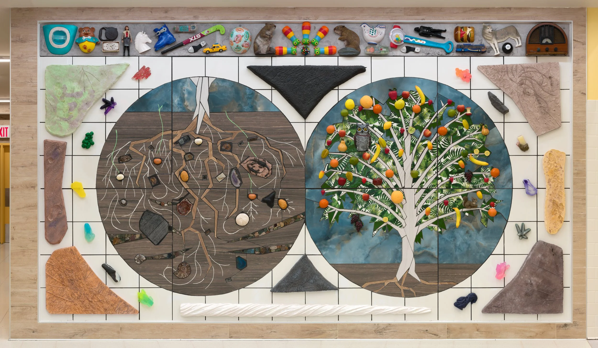 An overhead view of a large mosaic floor art piece depicting two trees, a barren tree on the left and a fruit-bearing tree on the right, crafted with various materials and surrounded by miscellaneous objects and rocks.