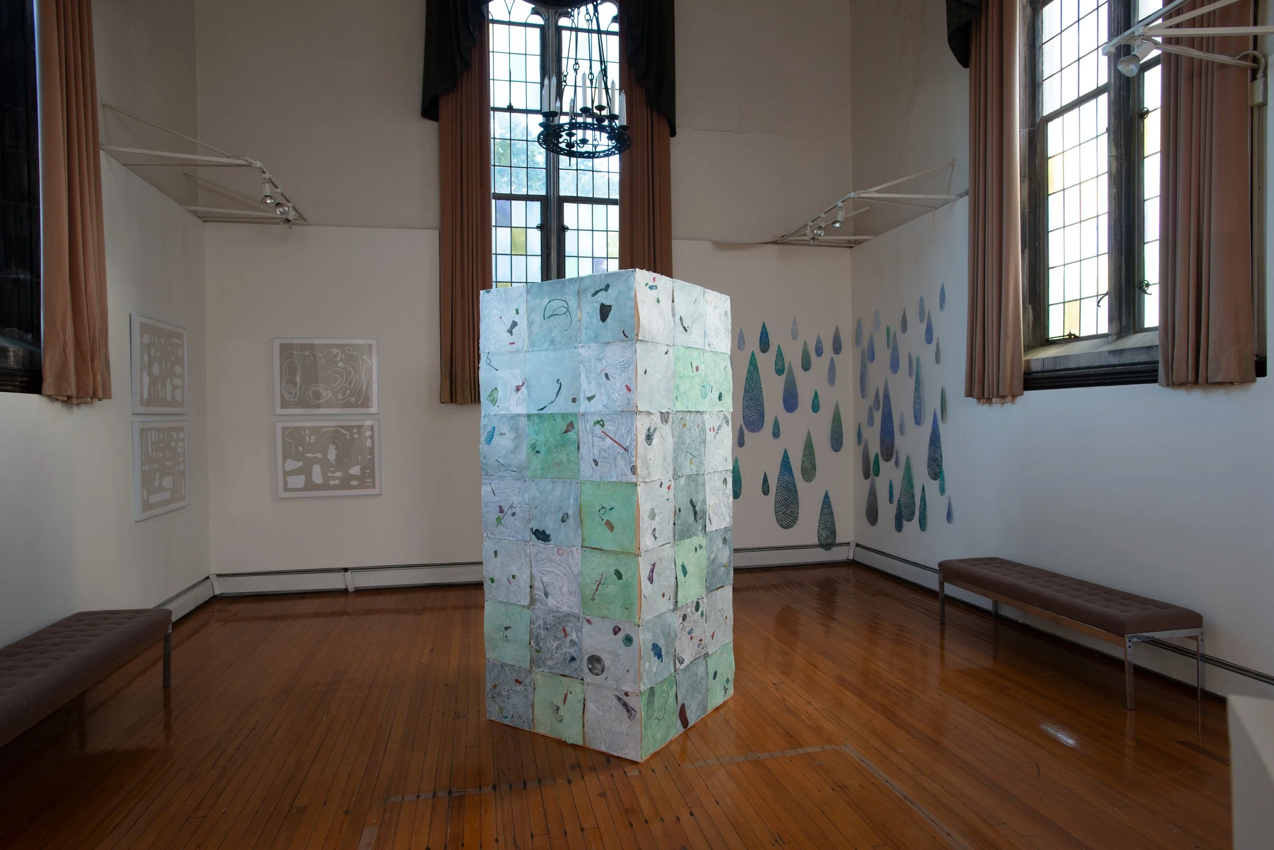 An art installation of a tall, rectangular structure made of small, colorful, textured paper or fabric squares in a gallery room. The room has wooden floors, large windows with brown curtains, and wall art including paintings and wall decoration.