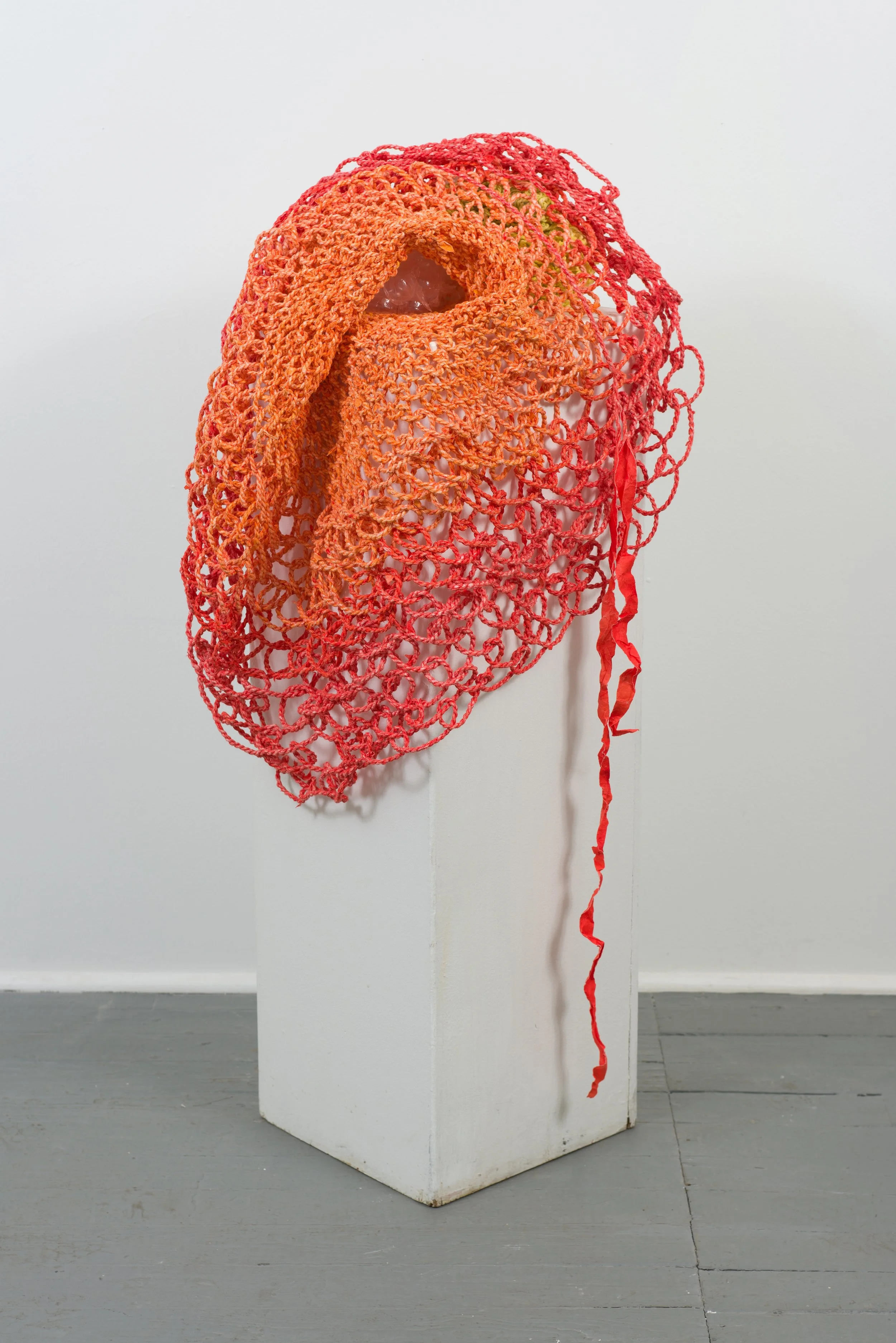 Mixed media sculpture with orange and red crochet-like netting draped over a glass object on a white pedestal.