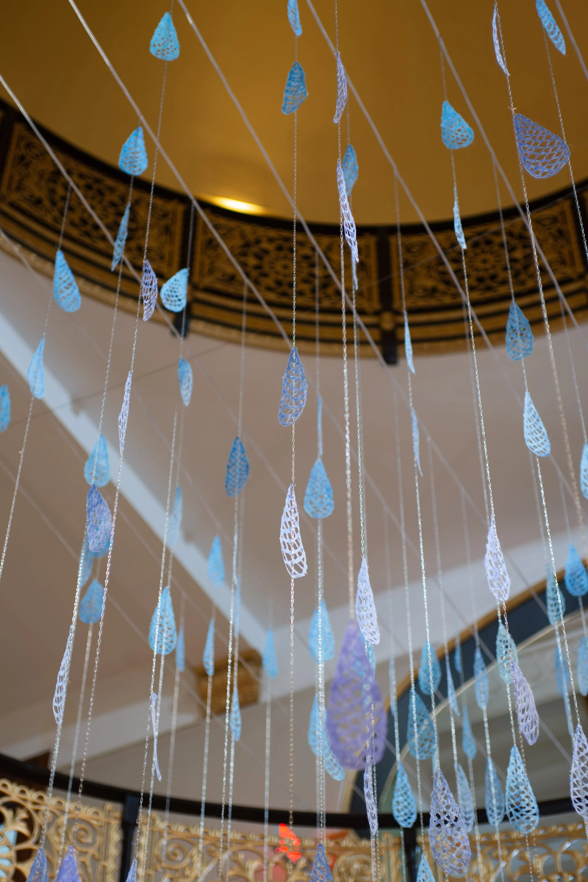 Decorative hanging art with blue teardrop-shaped cutouts hanging from strings, viewed from below inside a building with ornate gold railing and a domed ceiling.
