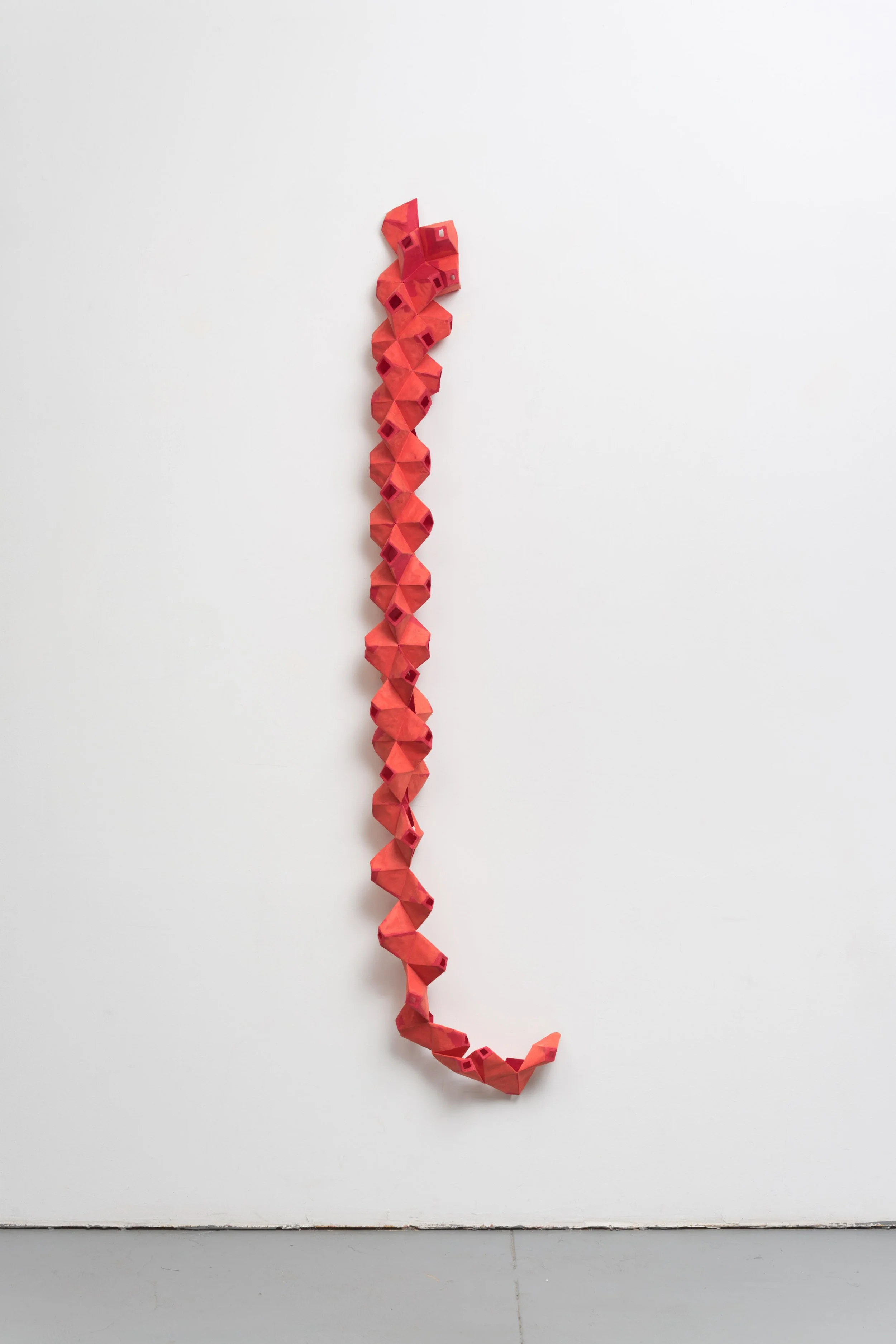 A white wall with a vertical chain of red paper or cloth geometric shapes hanging from top to bottom.