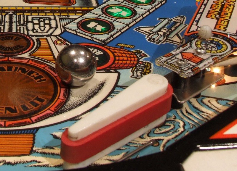 Close-up of a pinball machine playfield showing a silver ball, bumpers, and colorful artwork, including a spaceship and laser target symbols.