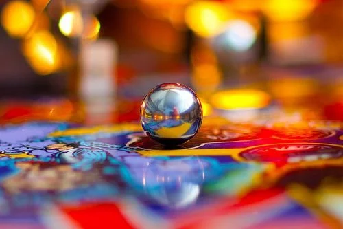 Close-up of a metal ball on a colorful pinball game surface with bright, blurry lights in the background.