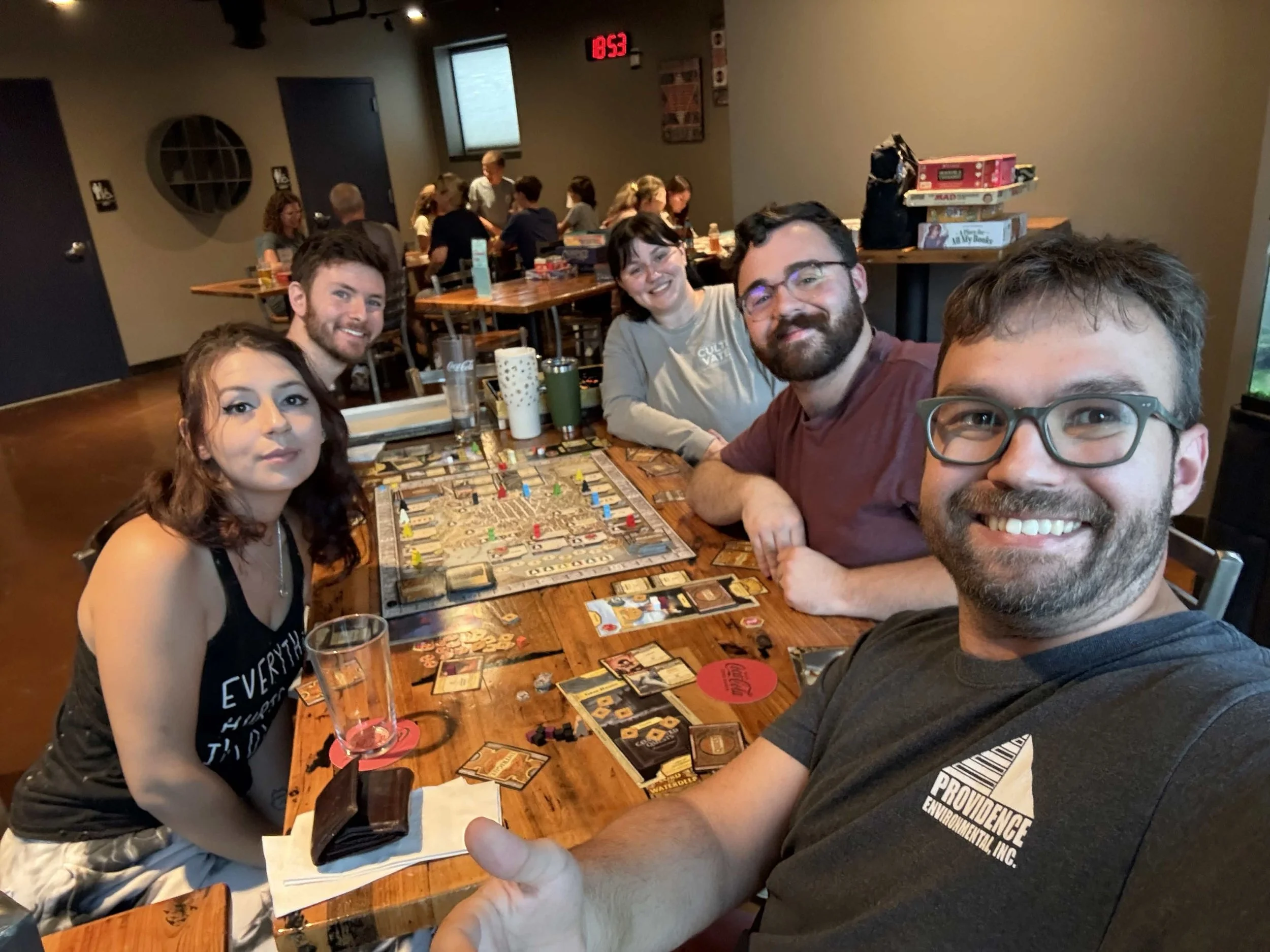 Five people smiling for a selfie around a wooden pub table covered in a board game, cards, and drinks, with a busy game night crowd visible at tables in the background.