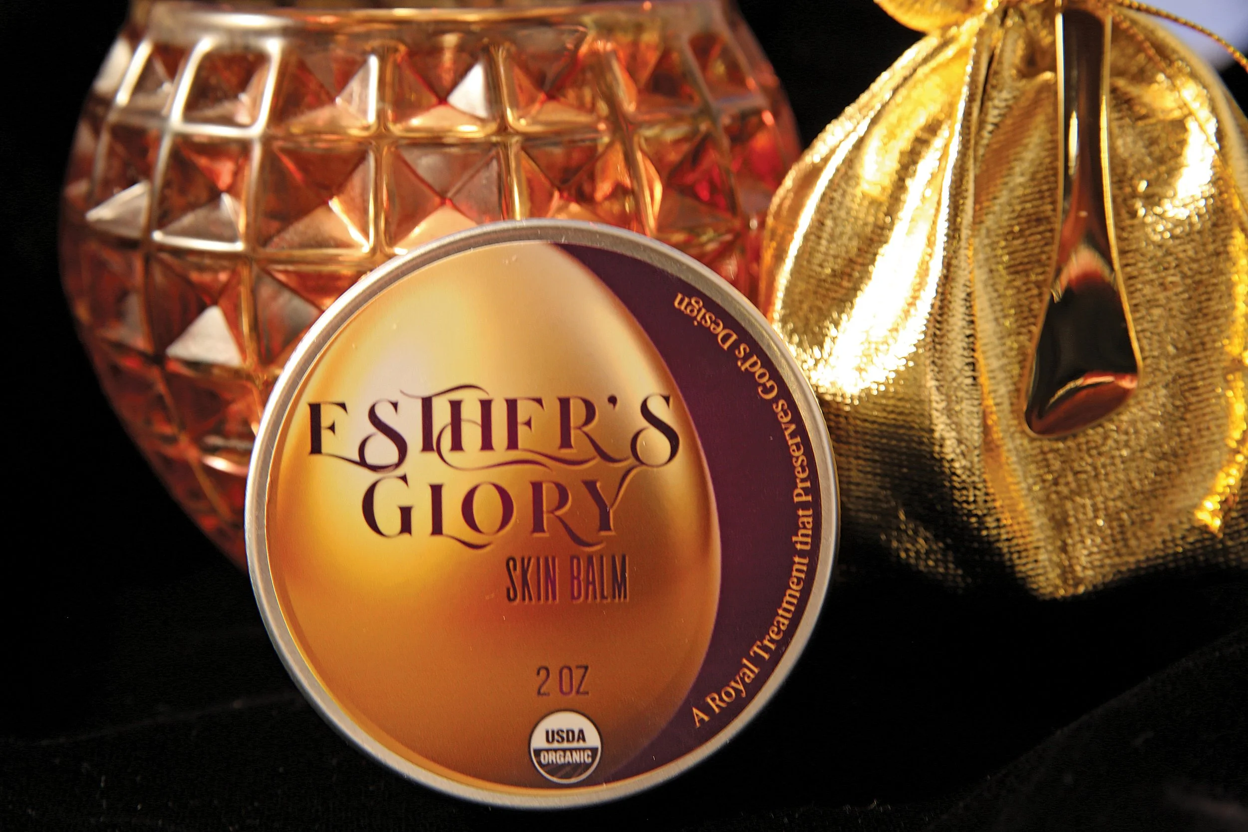 A container of Esther's Glory Skin Balm with gold decorative objects around it, including a textured pink and gold pumpkin and a shiny gold bag.