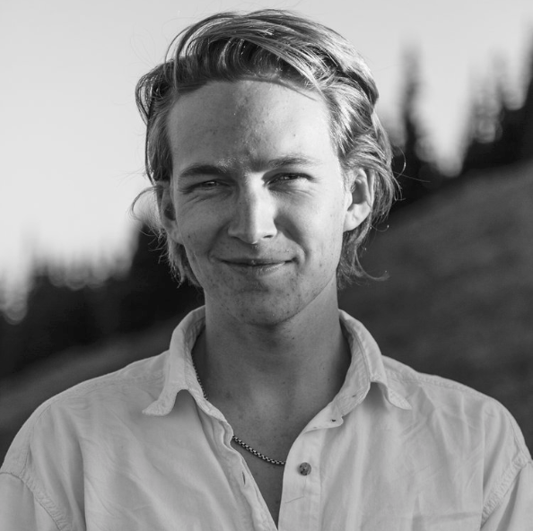 Black and white photo of a young man with light hair, wearing a collared shirt and a necklace, outdoors with blurred trees and hillside in the background.