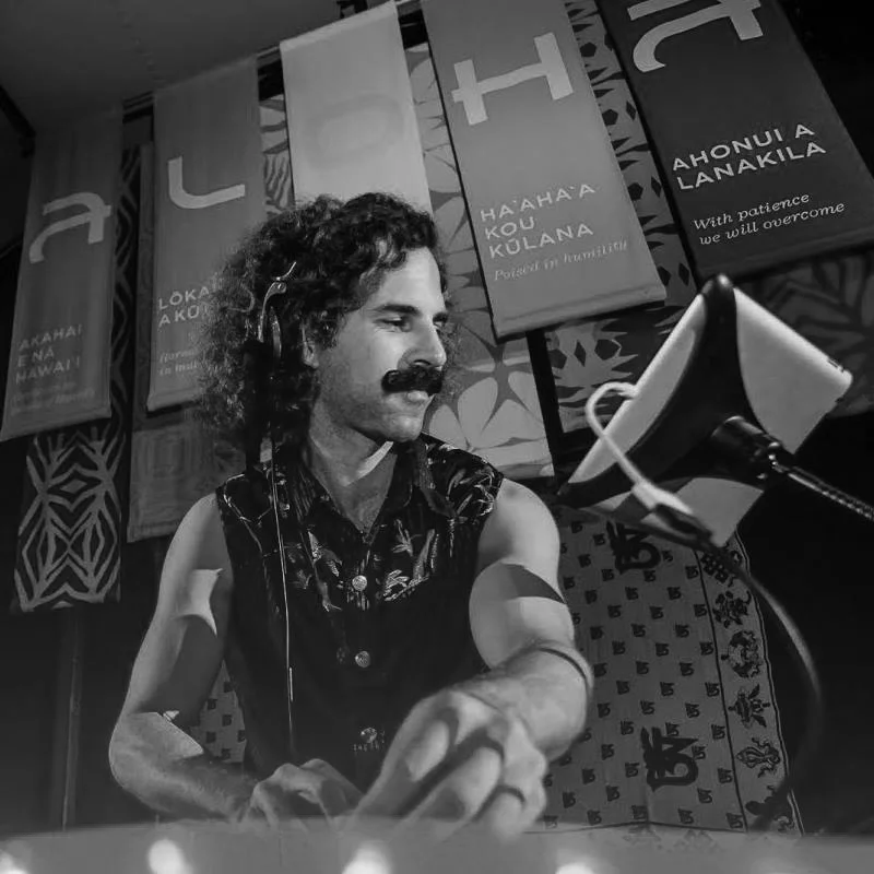 A man with curly hair and a mustache wearing headphones, operating music equipment in front of banners with Māori language phrases and designs.
