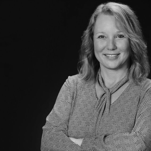 Black and white portrait of a smiling woman with shoulder-length wavy hair, wearing a patterned blouse with a bow tie at the neck, standing against a dark background.
