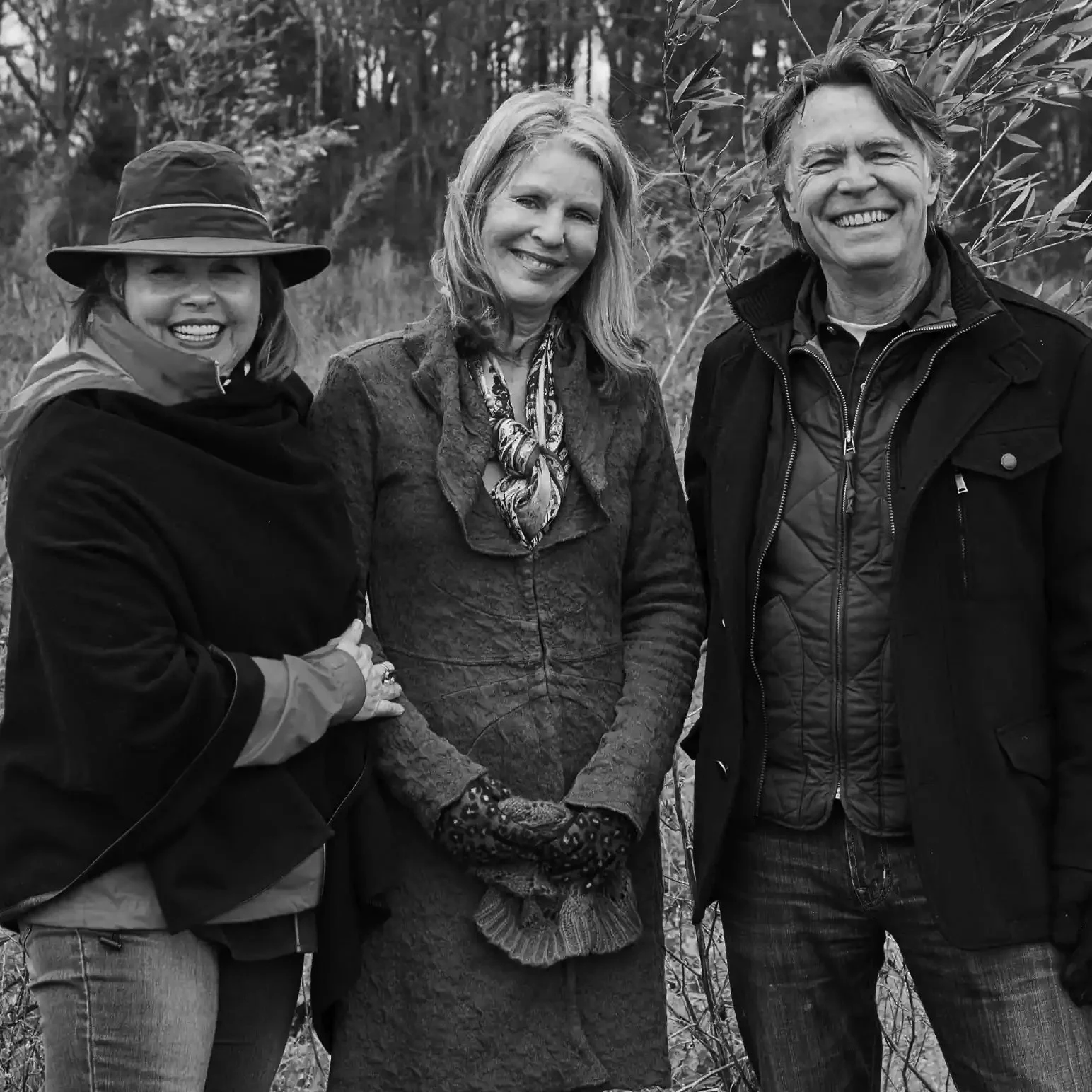 Three people standing outdoors among trees, smiling at the camera, in black and white.