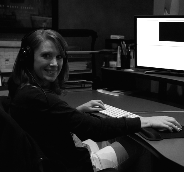 A woman with headphones sitting at a desk in a dark office, smiling at the camera, with a computer monitor and office supplies in the background.