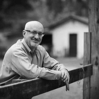 Black and white photo of smiling man with glasses leaning on a wooden fence outdoors, with a house in the background.